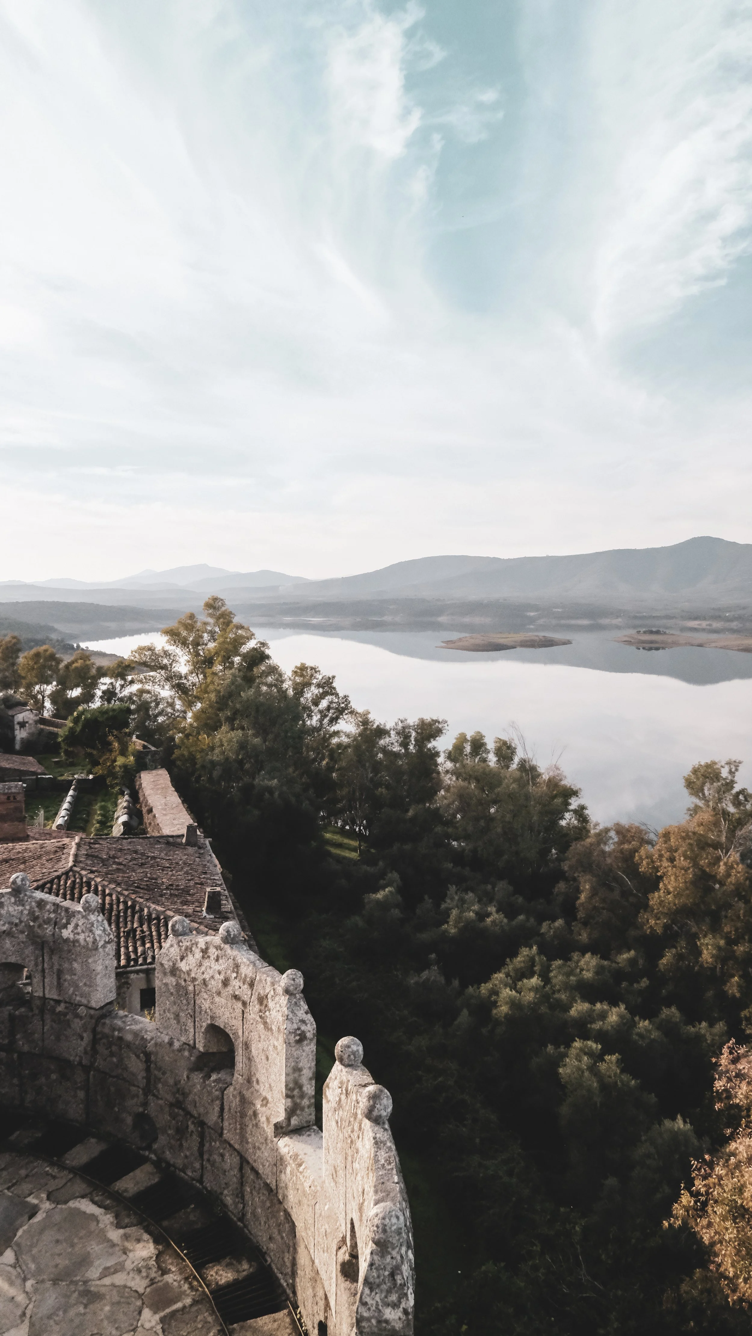 A scenic view of a lake with mountains in the background, trees in the foreground, and a stone or concrete railing along a pathway.