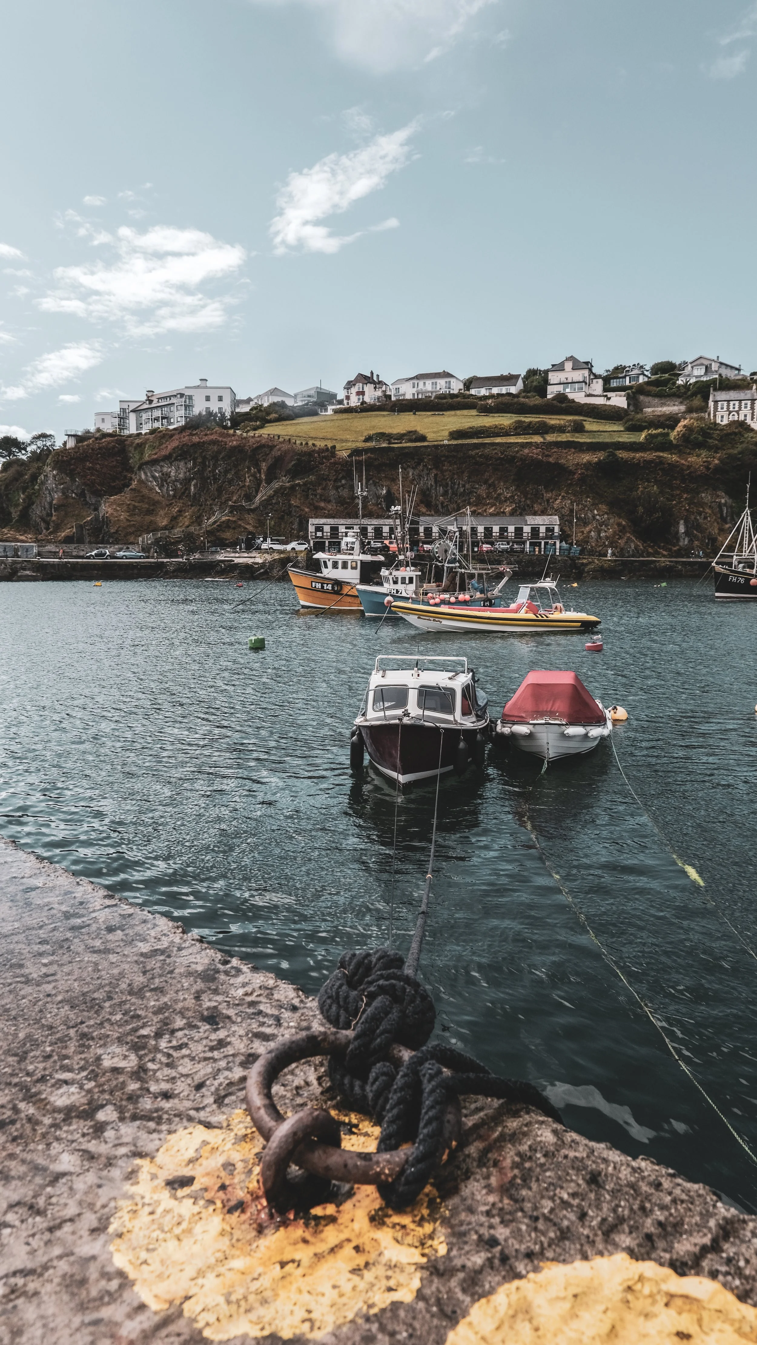 Boats docked at a harbor with houses on a hillside in the background under a partly cloudy sky.