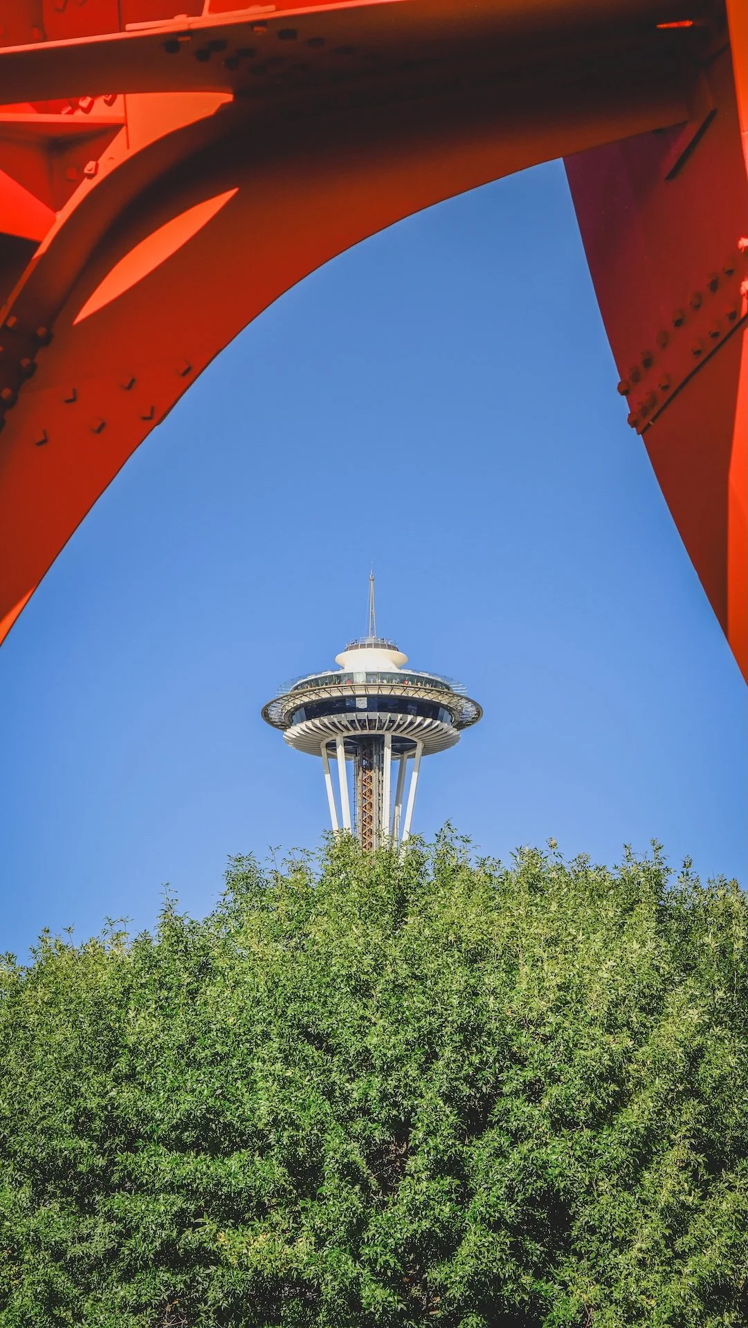 The Space Needle, a tall observation tower, seen from below with trees at the base and a blue sky background, framed by a red structural element.