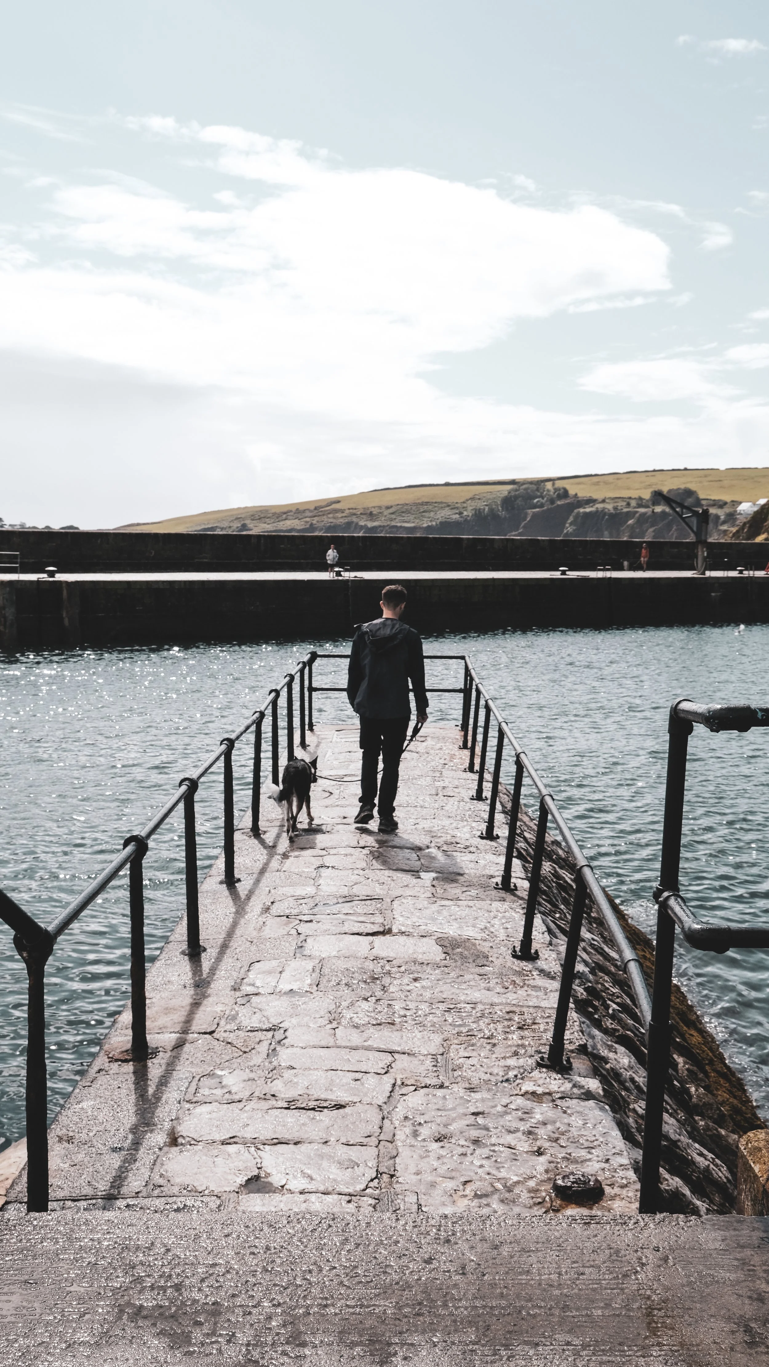A man walking his dog on a stone pier over water, with hills and a cloudy sky in the background.