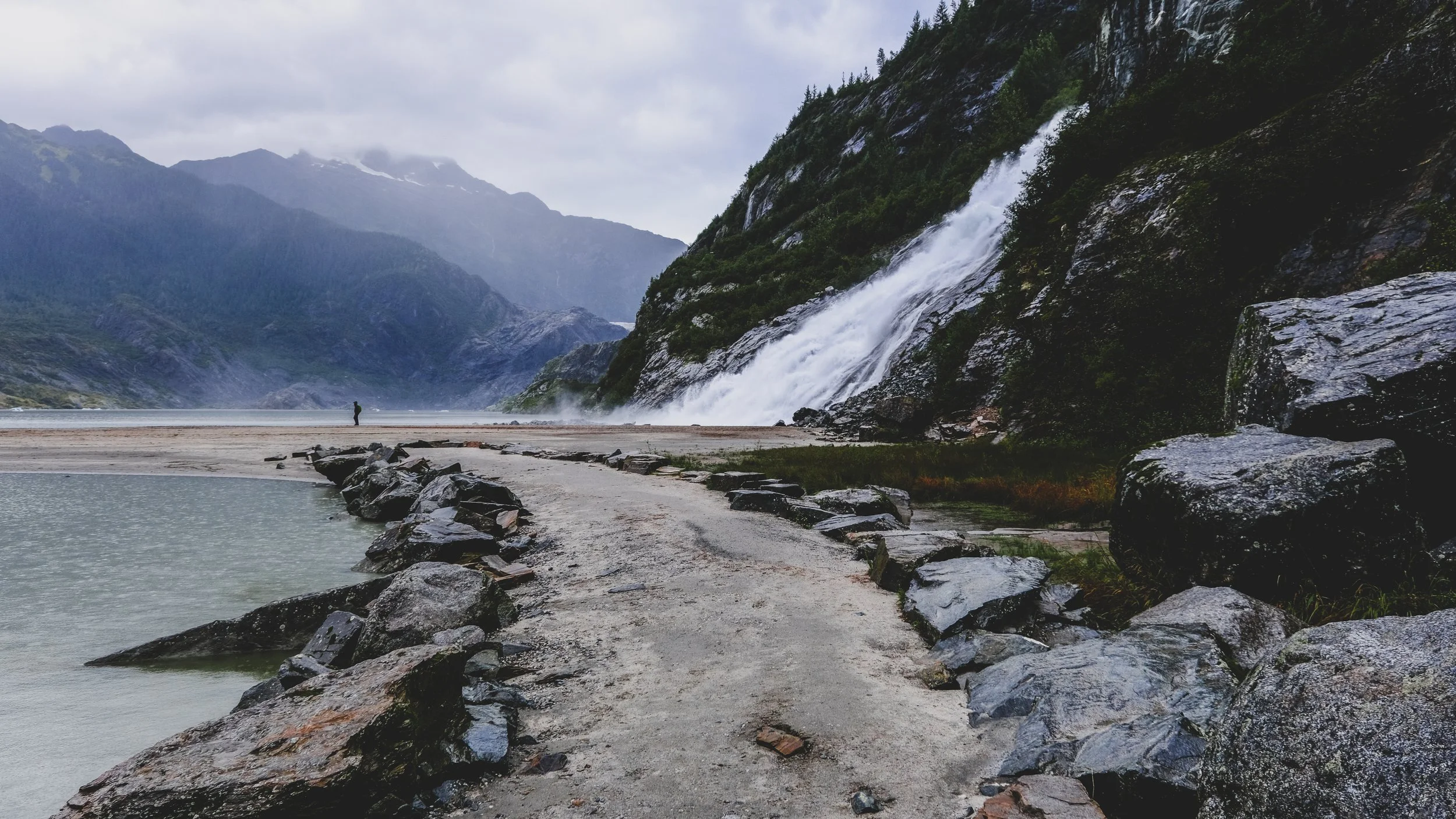 A rugged beach with large rocks, a dirt path, a person in the distance, and a waterfall cascading down a mountain on a cloudy day.