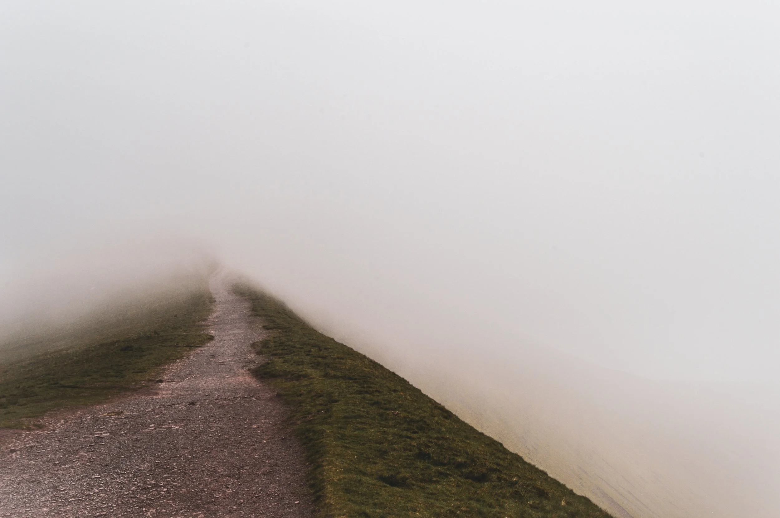 A narrow dirt and gravel trail on a grassy hill, disappearing into thick fog.