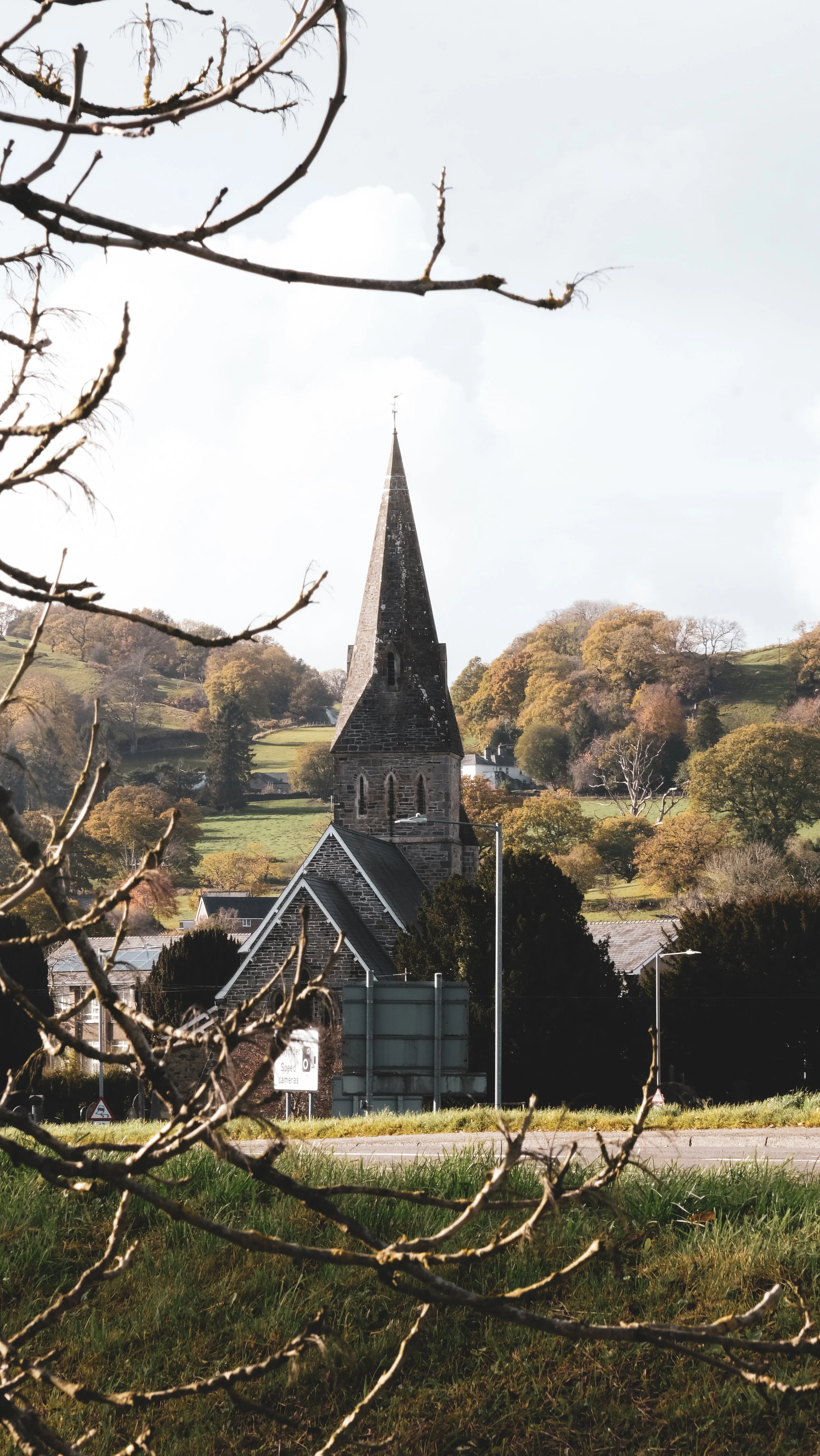 A stone church with a tall steeple, surrounded by trees, on a hillside under a cloudy sky.