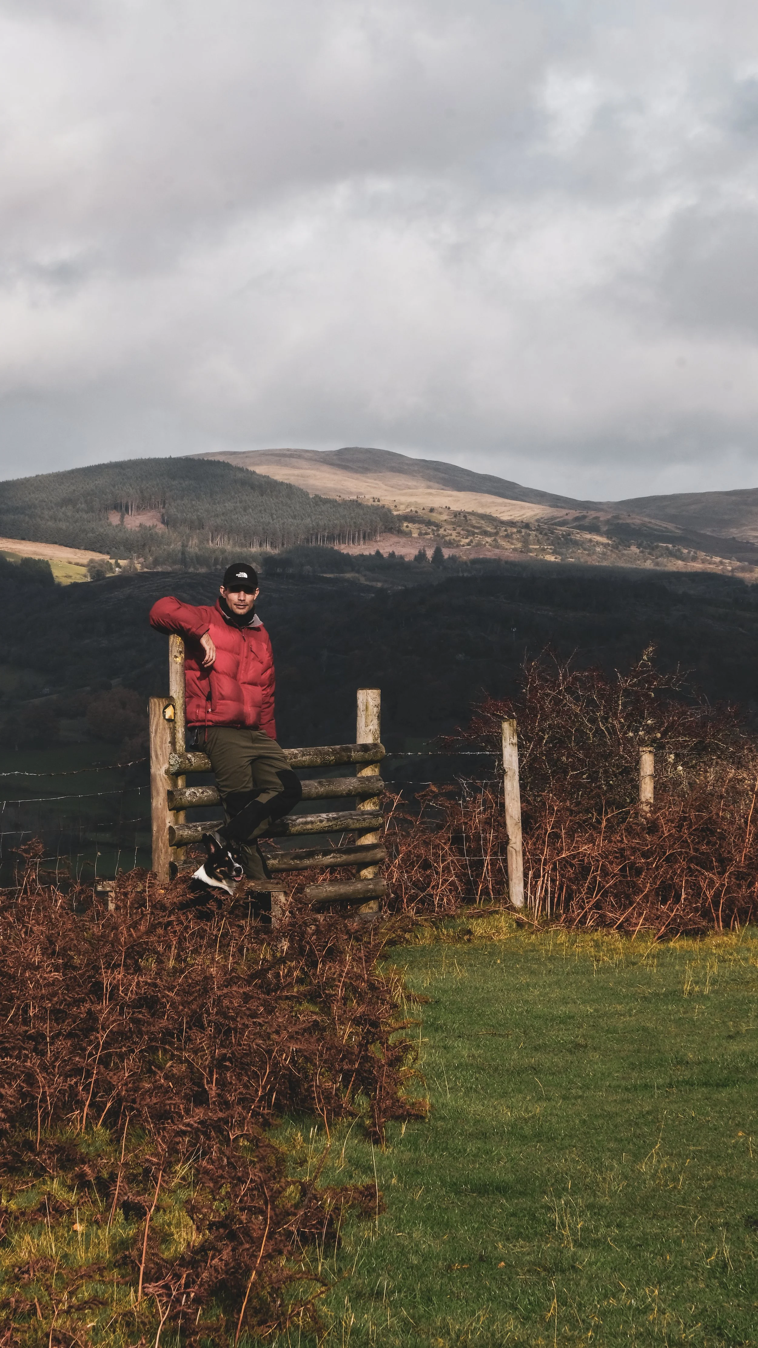 A man in a red jacket and black cap sitting on a split-rail wooden fence in a countryside landscape with hills and cloudy sky in the background, accompanied by a small black and white dog.