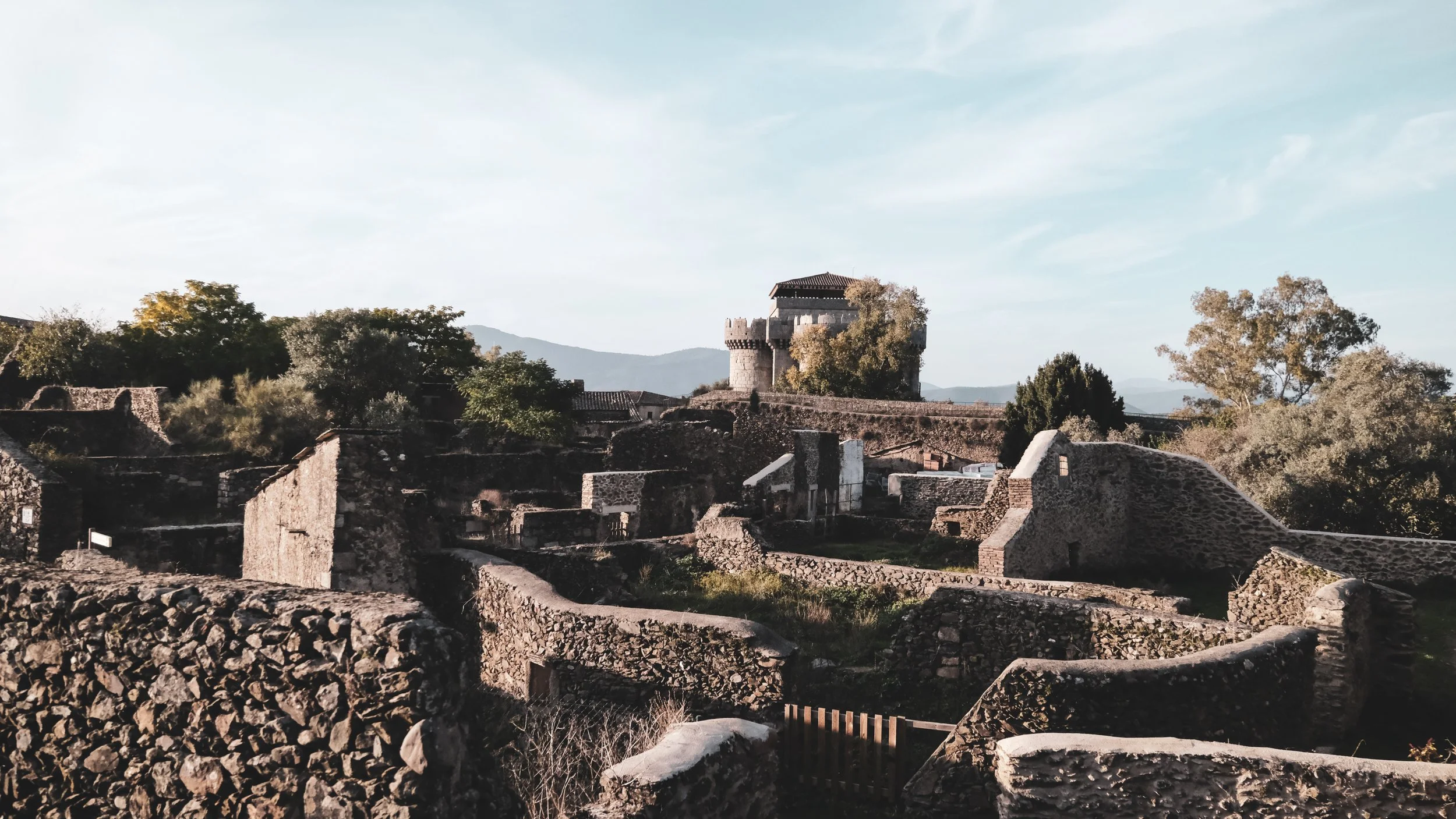 Ancient stone ruins with a castle tower in the background, surrounded by trees and mountains, under a partly cloudy sky.
