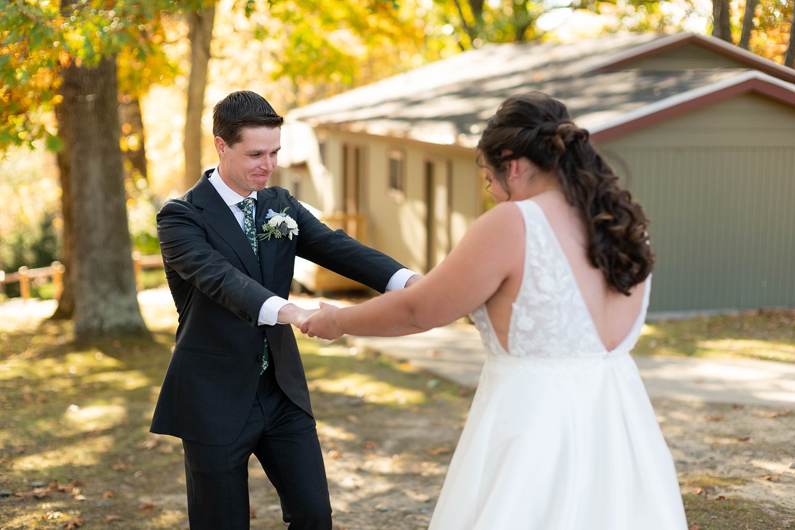 A groom in a dark suit and a bride in a white dress holding hands outdoors, surrounded by trees and a cabin.