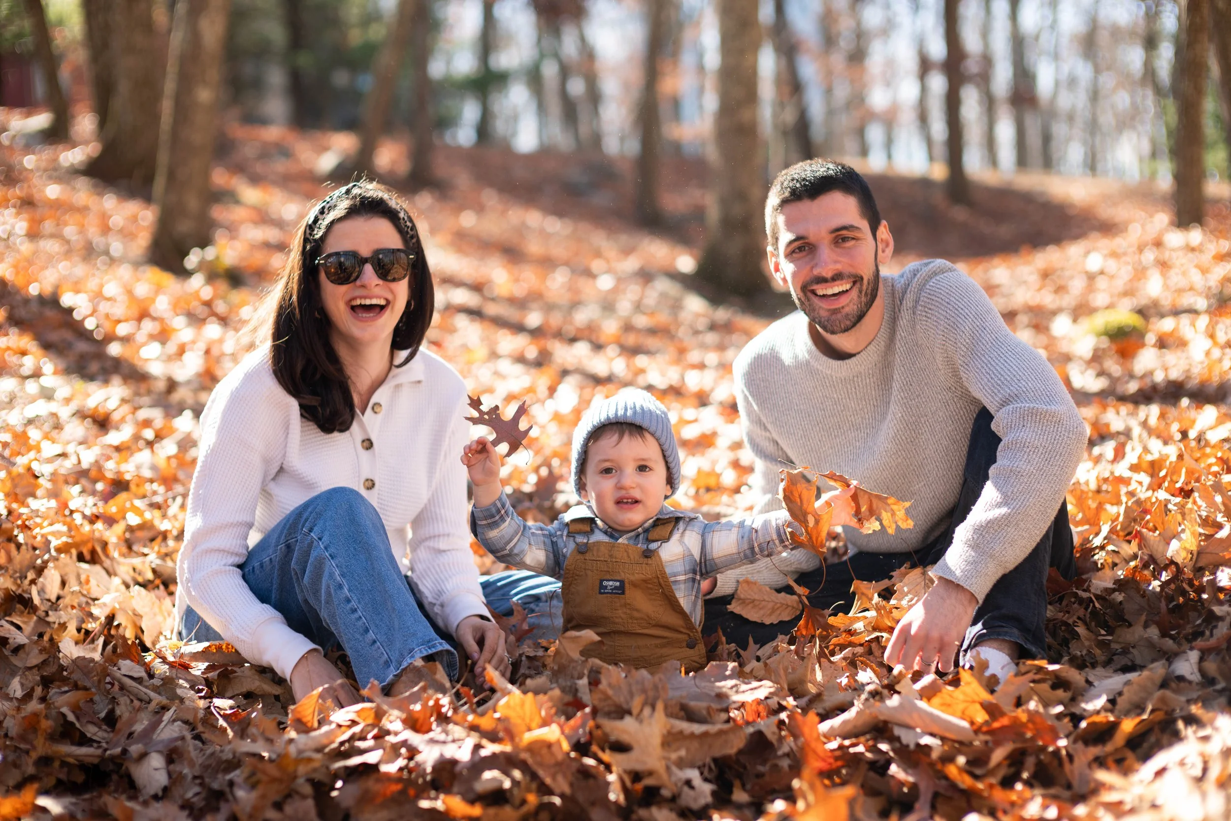 Family sitting in autumn leaves in a forest, smiling and playing with their child.