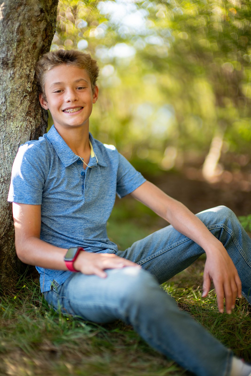 A young boy with braces sitting outdoors against a tree, wearing a blue polo shirt and jeans.