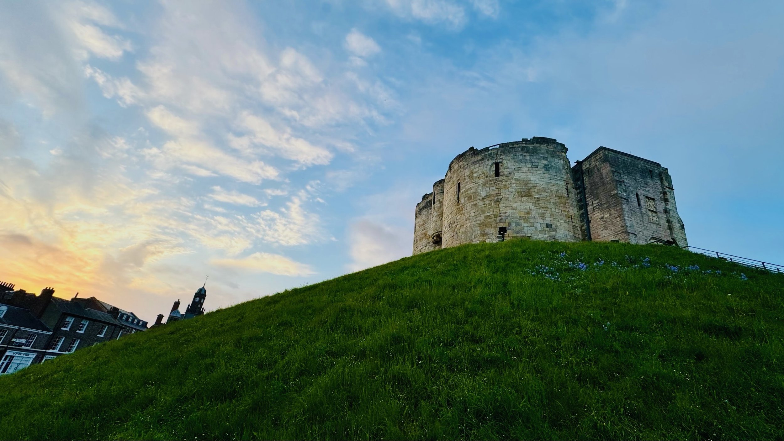 Stone castle on a grassy hill with a tower in the background under a sunset sky.