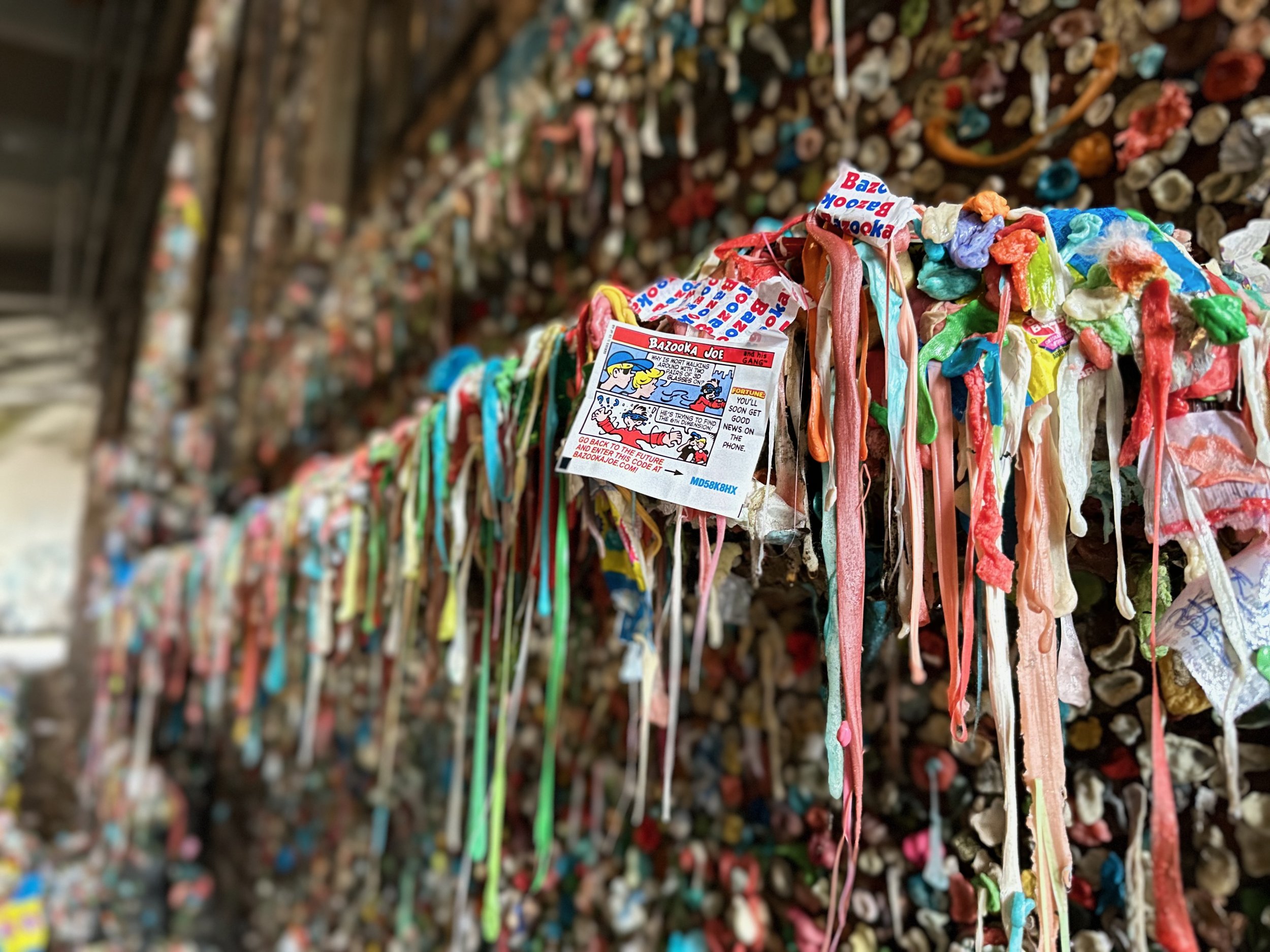 Close-up of a wall covered in colorful chewing gum, with Bazooka Joe gum wrappers visible among the gum pieces.