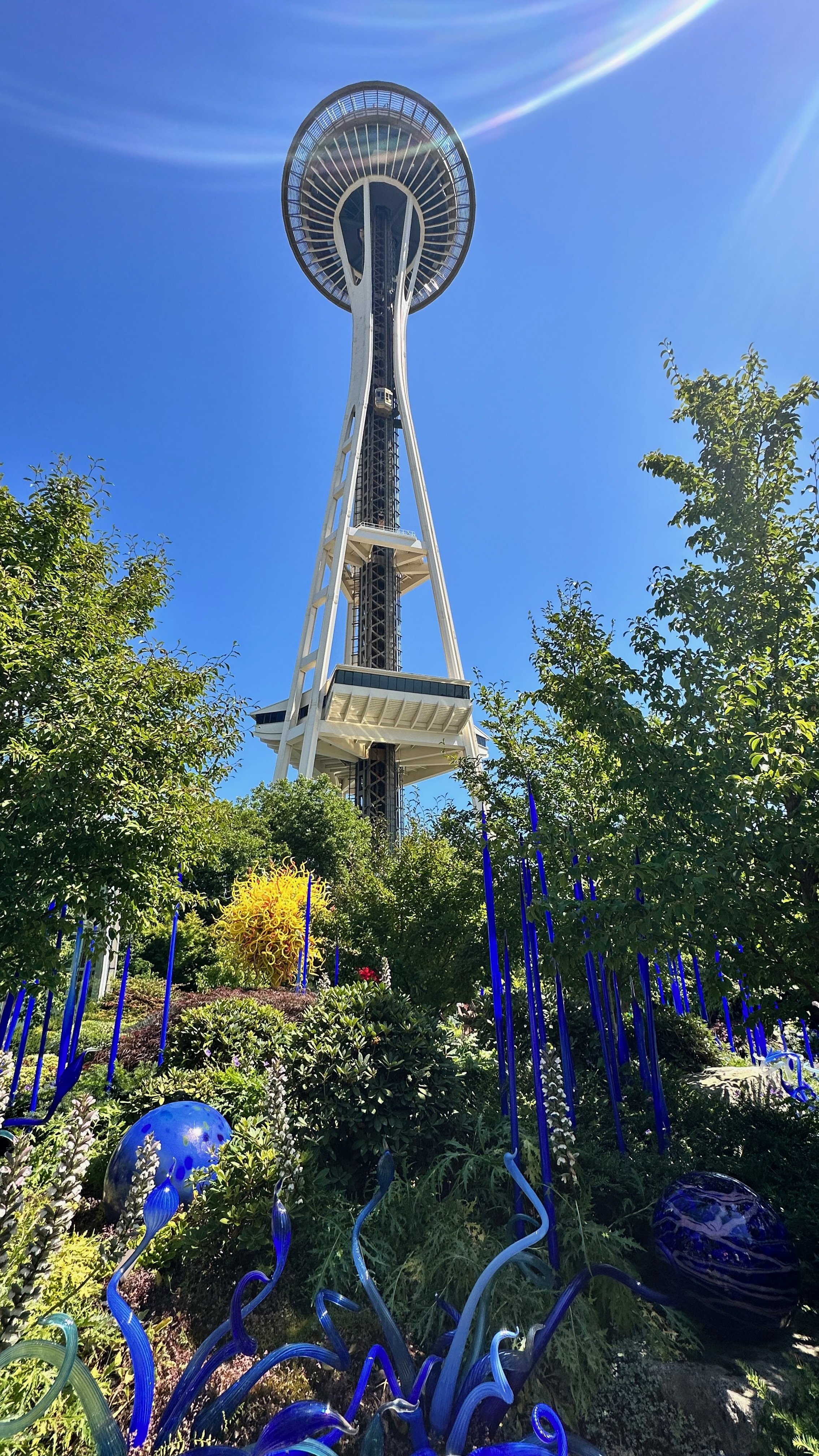 Space Needle in Seattle with Chihuly Garden and Glass artwork, blue sculptures, and green foliage on a sunny day.