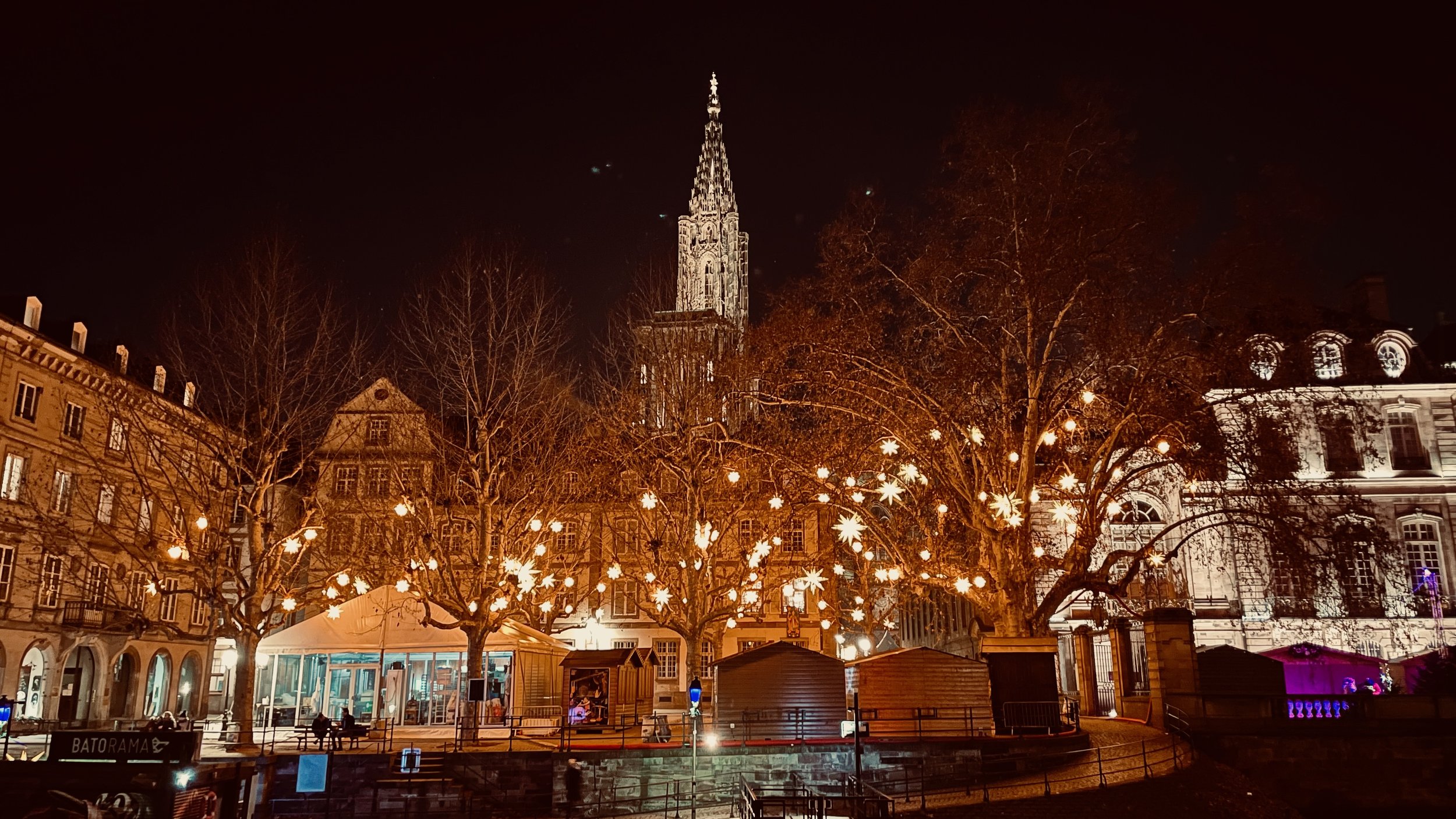 Night scene in Strasbourg, France with illuminated buildings, a cathedral spire, holiday lights in trees, and a festive market setup.