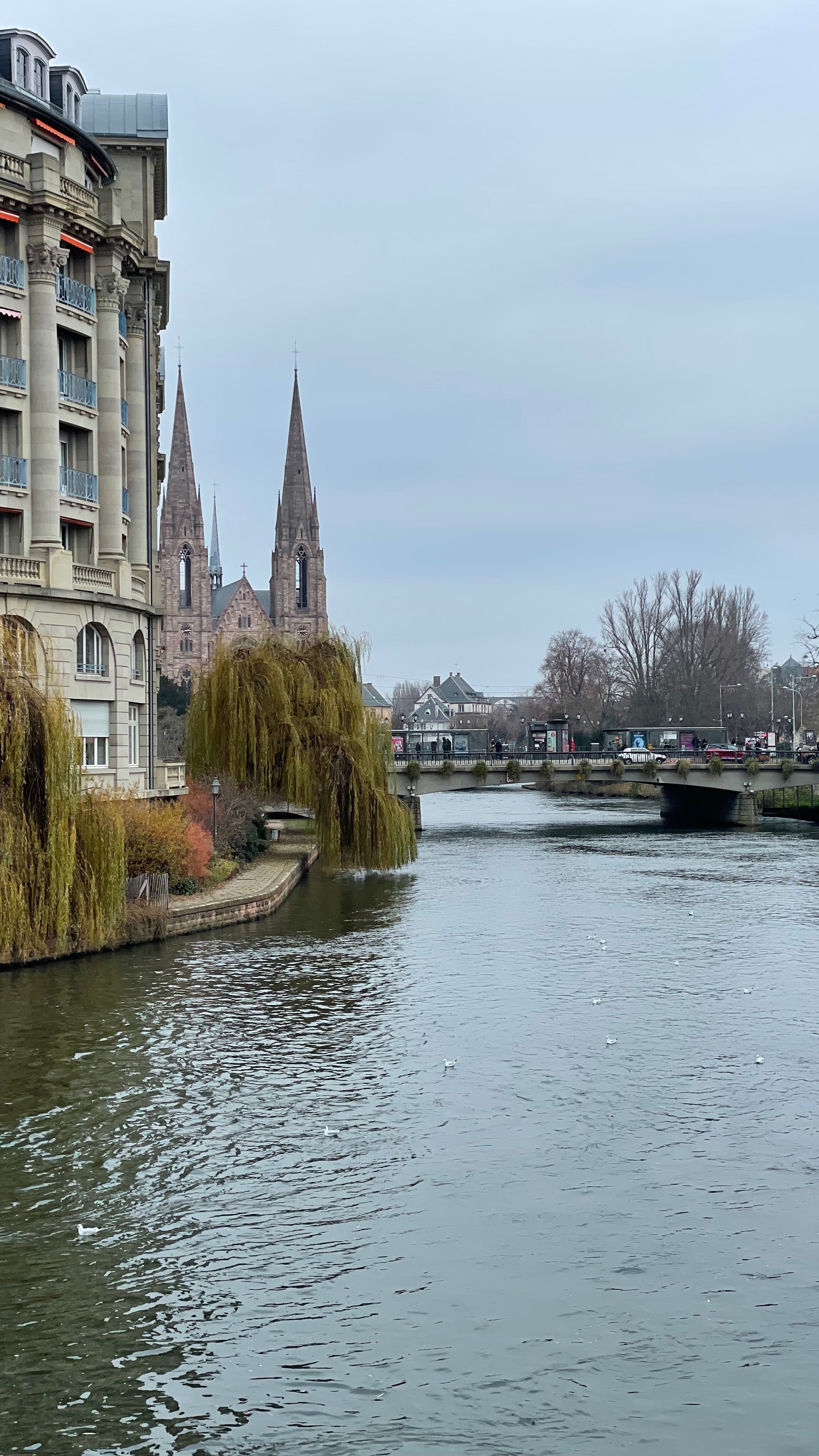 River scene with a building on the left, willow trees, a bridge, and church spires in the background on a cloudy day.