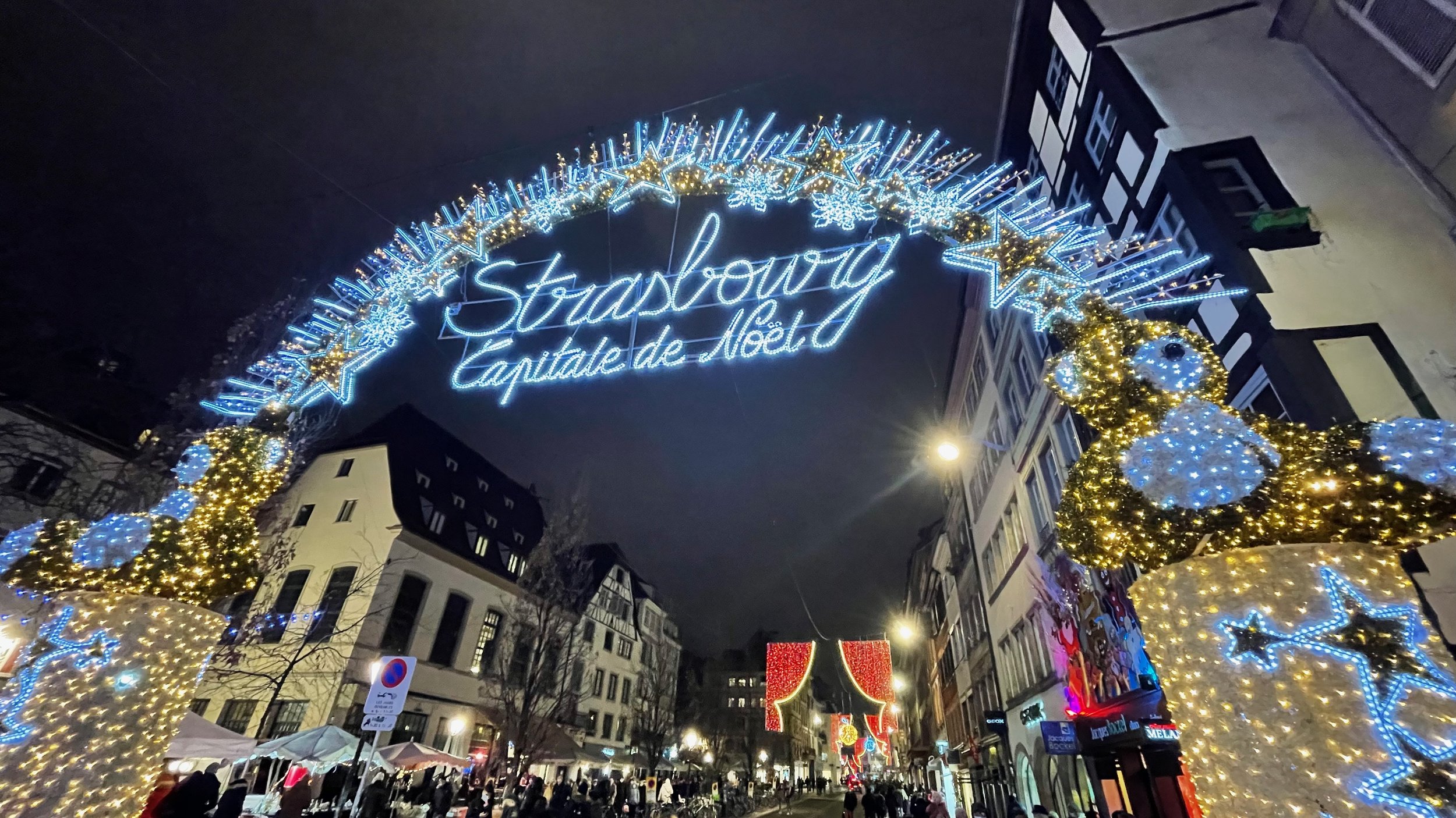 Christmas lights display reading 'Strasbourg Capitale de Noël' in an illuminated archway at night on a city street with festive decorations and buildings.