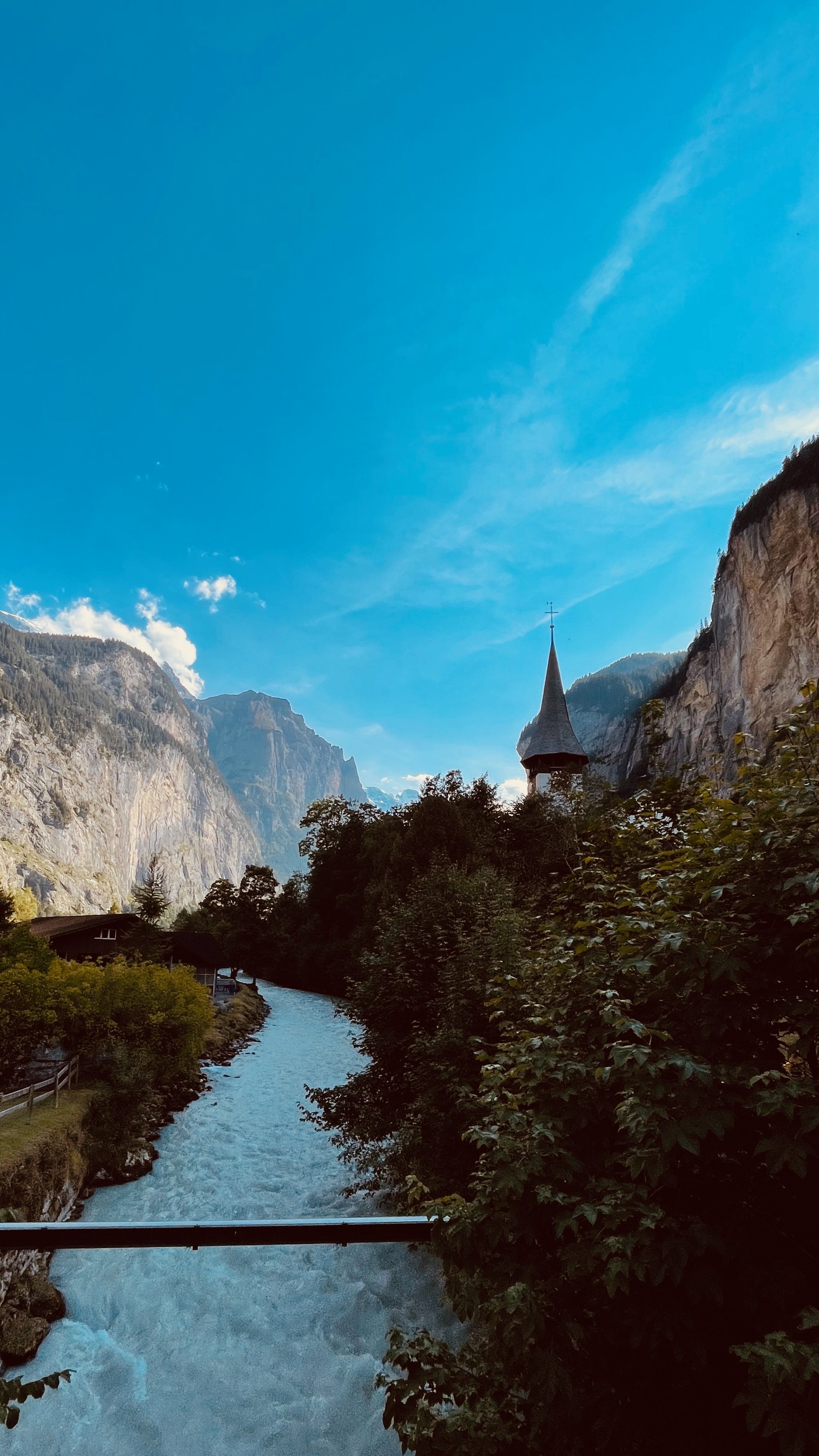 Scenic landscape with a flowing river, surrounded by lush greenery and cliffs, beneath a bright blue sky. A church steeple is visible among the foliage.