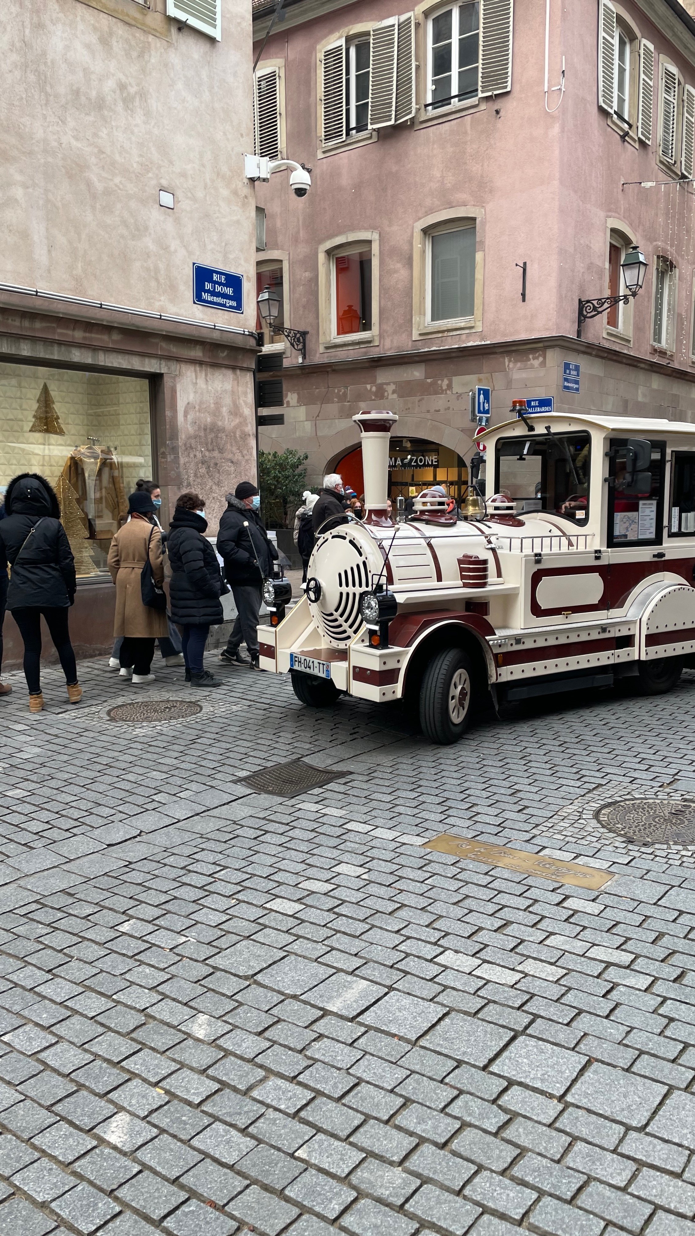 A small tourist train with a locomotive design parked on a cobblestone street in a European city. People wearing winter clothing and masks are nearby, and buildings with shuttered windows are in the background. A street sign reads 'Rue du Dôme.'