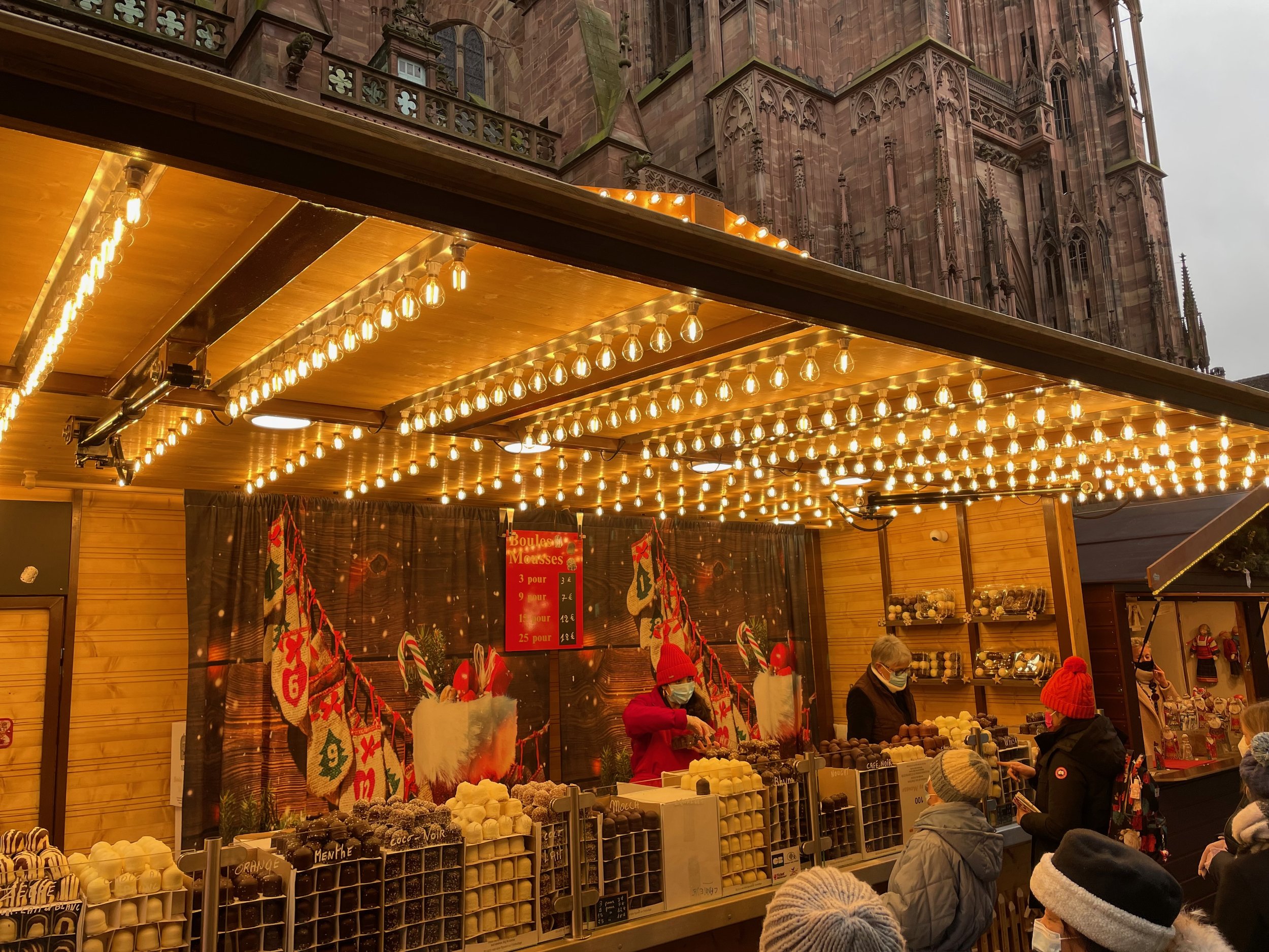 Christmas market stall selling chocolates and festive goods under string lights, with people shopping and a historic cathedral in the background.