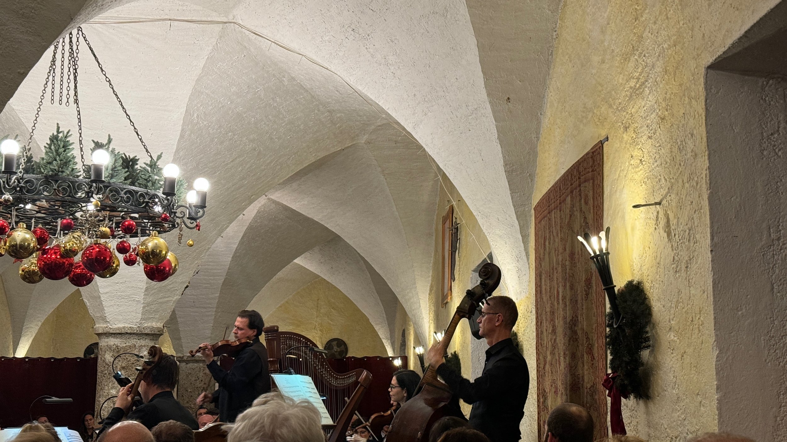 Orchestra performing in elegant venue with musicians playing string instruments, decorated chandelier with gold and red ornaments, arched ceiling, and warm lighting.