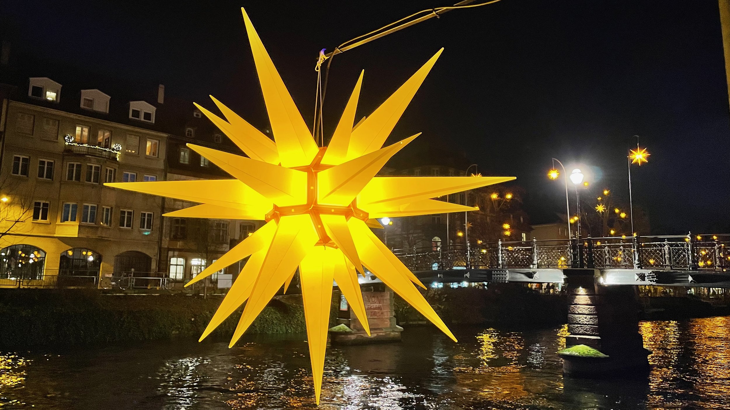Brightly lit yellow star decoration hanging over a river at night, with a bridge and buildings in the background.