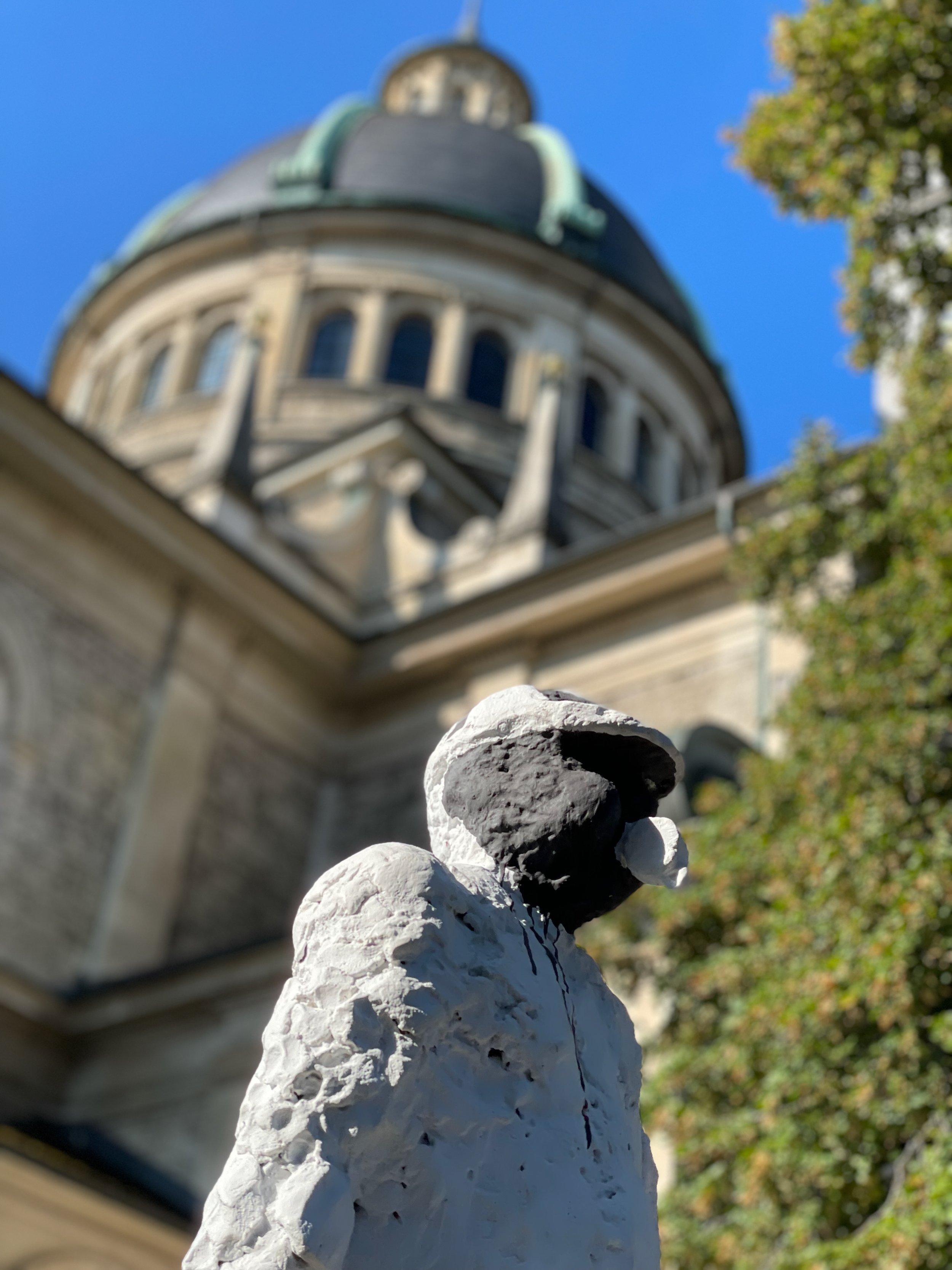 Sculpture of a bird in front of a historic domed building with trees and blue sky.