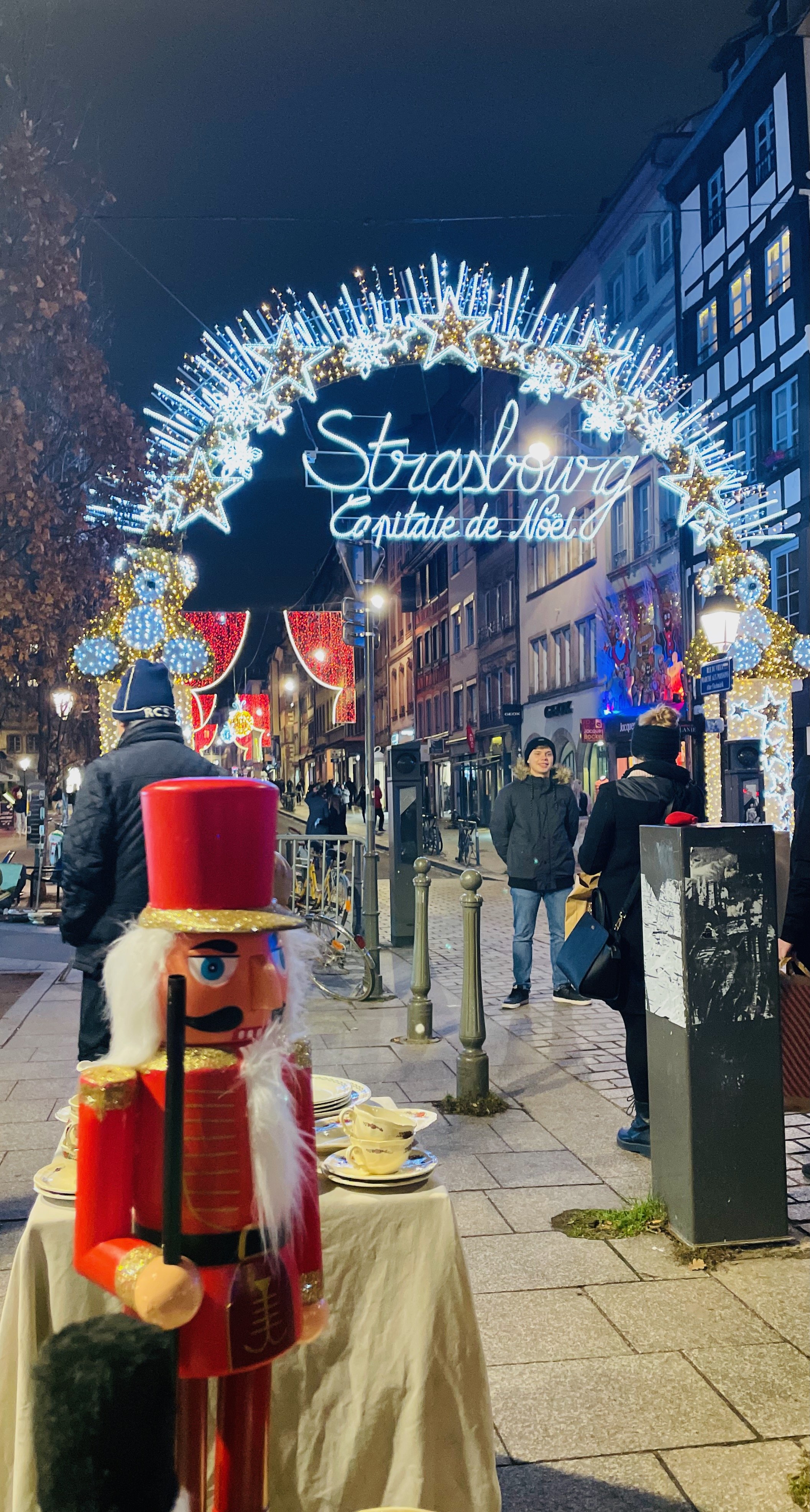 Christmas decorations in Strasbourg, featuring a Nutcracker, festive lights, and a sign reading 'Strasbourg Capitale de Noël' in a vibrant street scene.