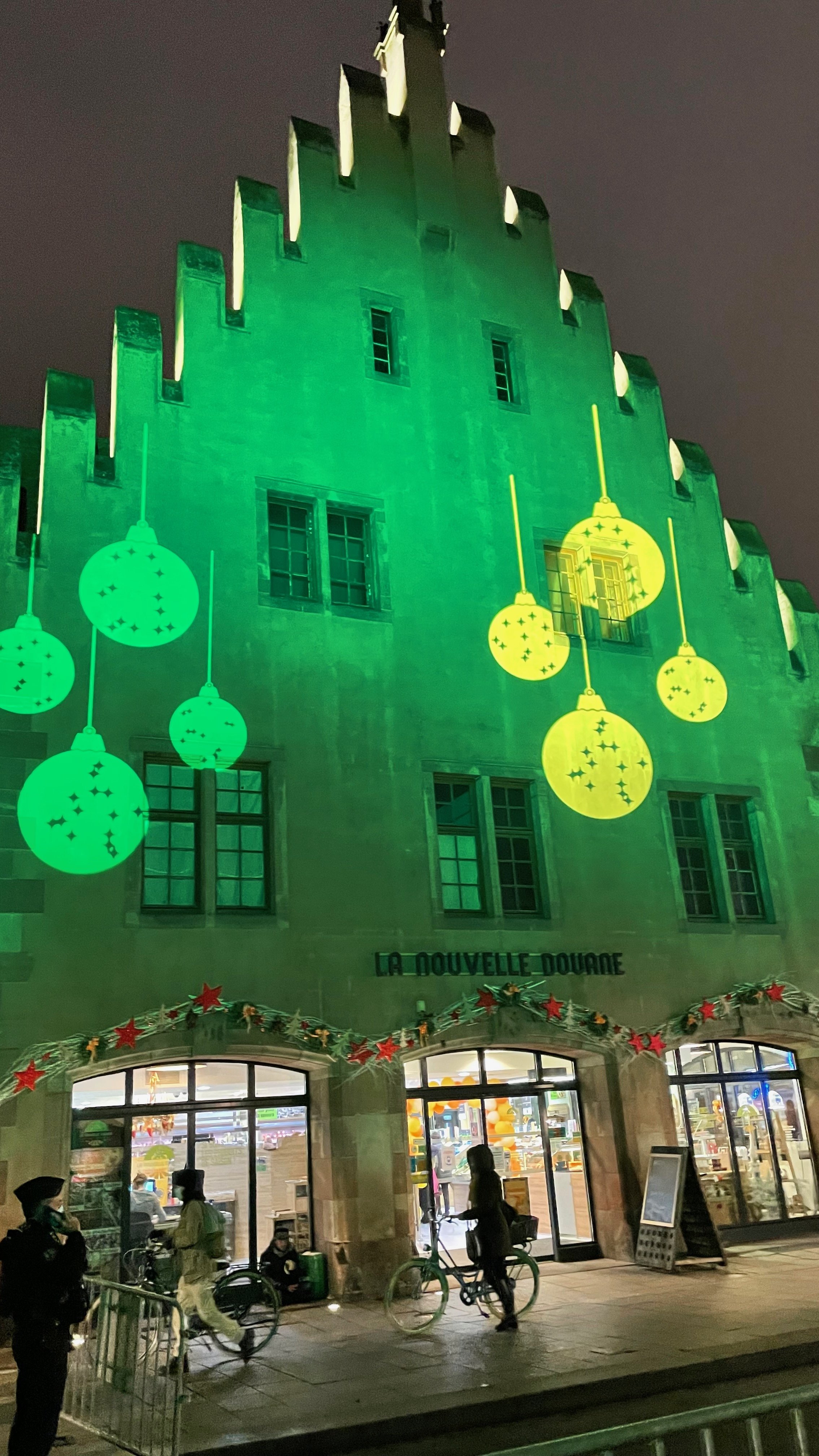 Holiday-themed lights projecting green and yellow ornaments on a building facade, "La Nouvelle Douane," with people passing by on bicycles, adorned with festive red star decorations.