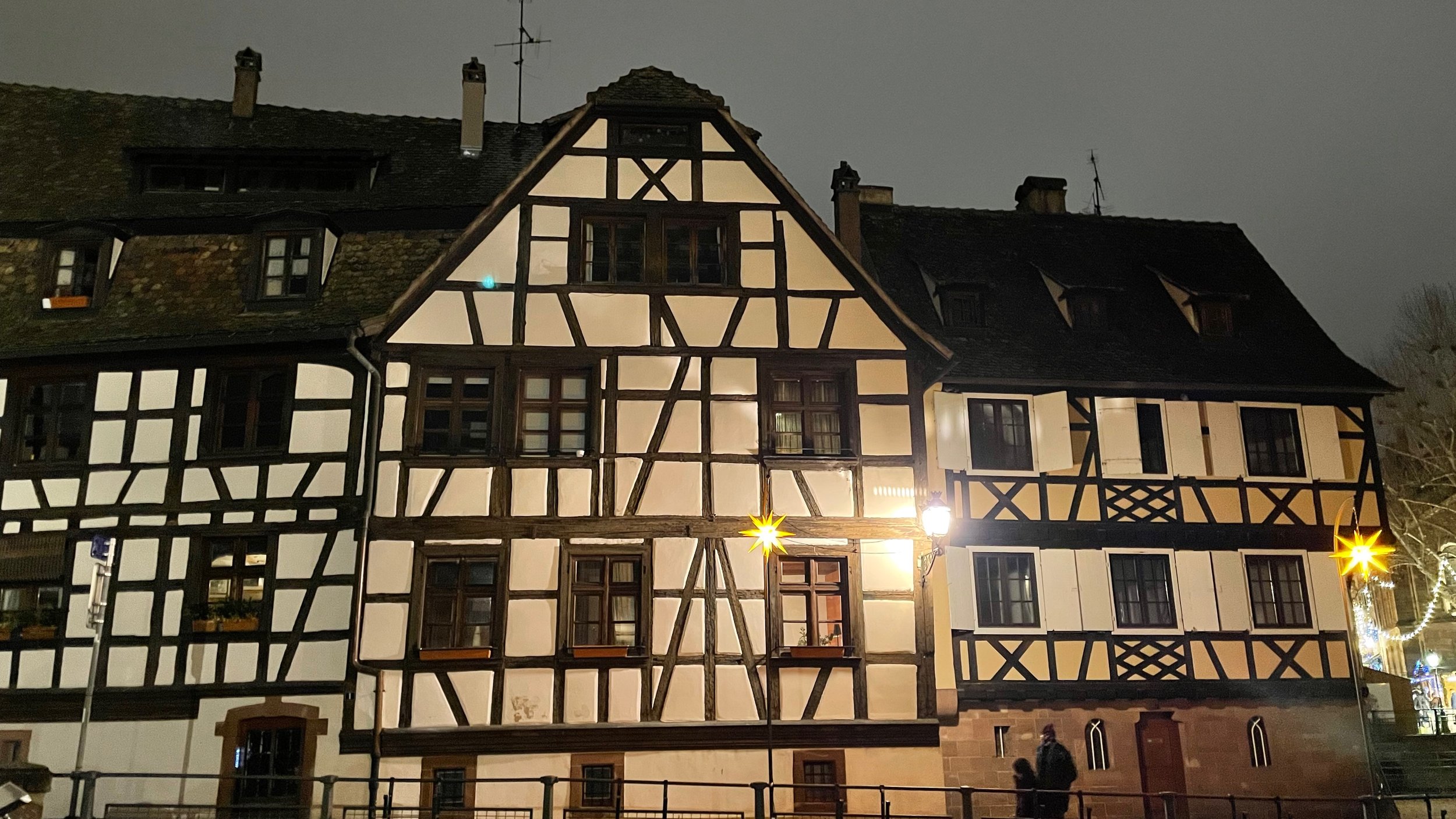 A traditional half-timbered house at night, featuring white walls with dark wooden beams, in Strasbourg, France. The building is illuminated, and a lamppost with a star-shaped decoration is visible in the foreground.