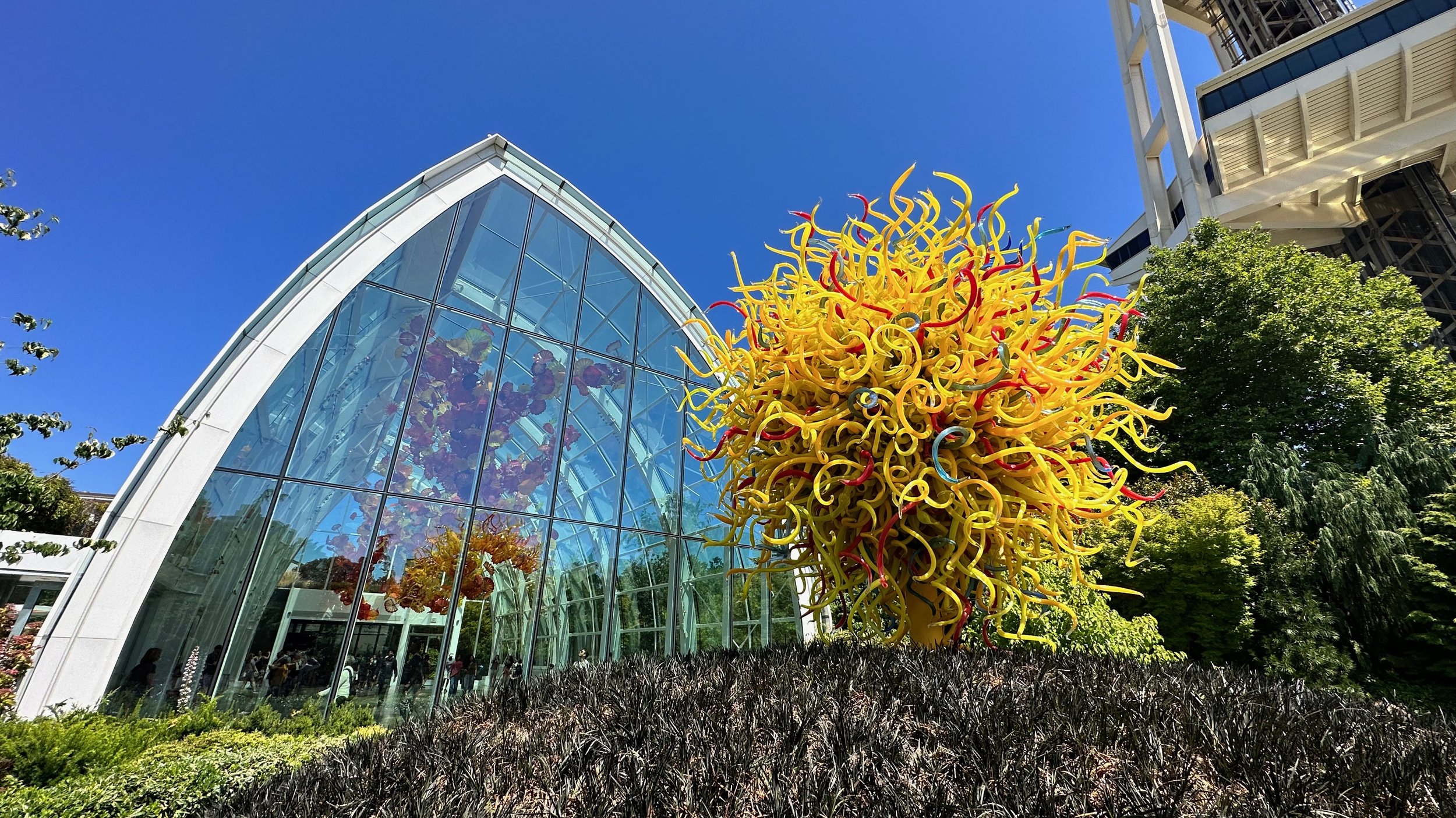 Glass art installation with colorful swirls and a large glass building with arched roof under a clear blue sky.