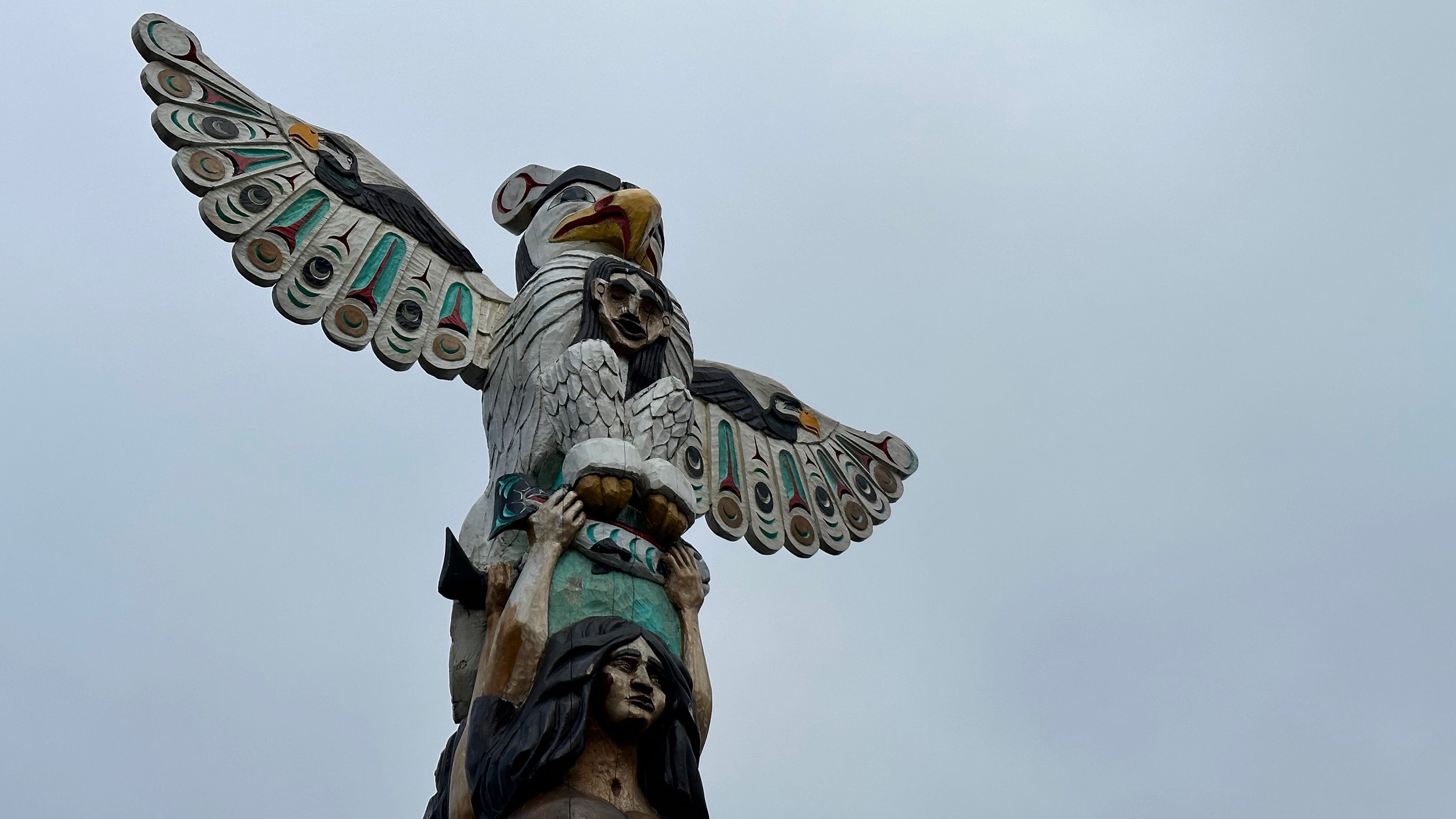 Close-up of a totem pole featuring an eagle with outstretched wings and a human figure below against a cloudy sky.
