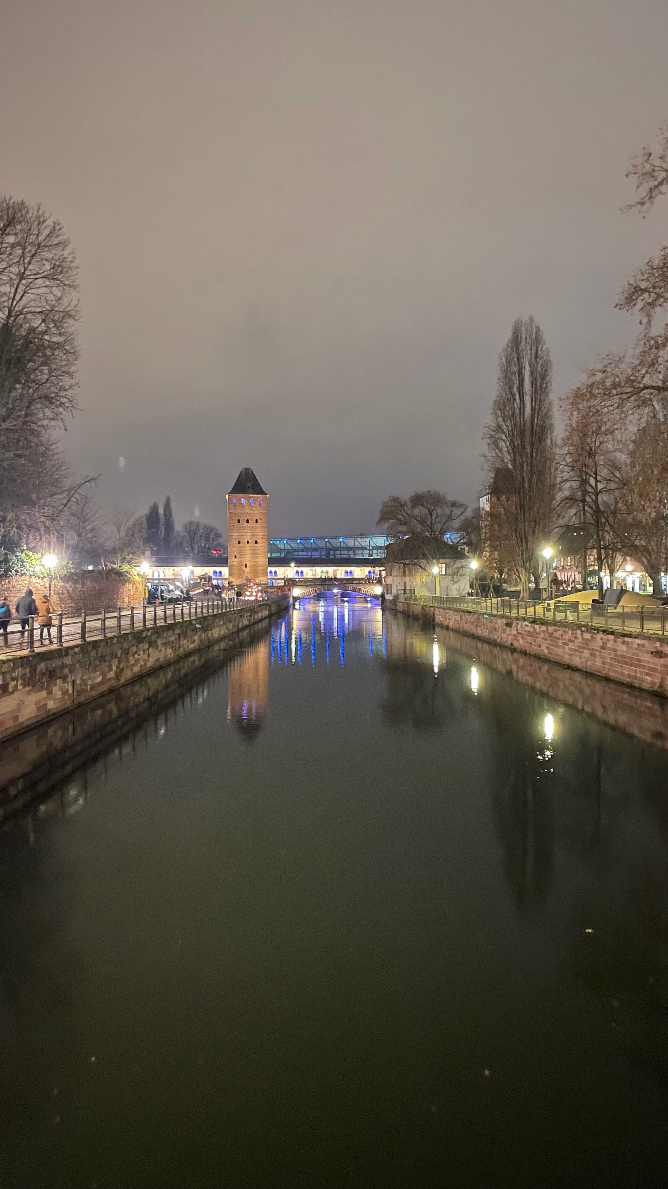 Evening view of a canal flanked by trees and stone walls, with a stone tower and a bridge in the background, illuminated by streetlights and reflections in the water.