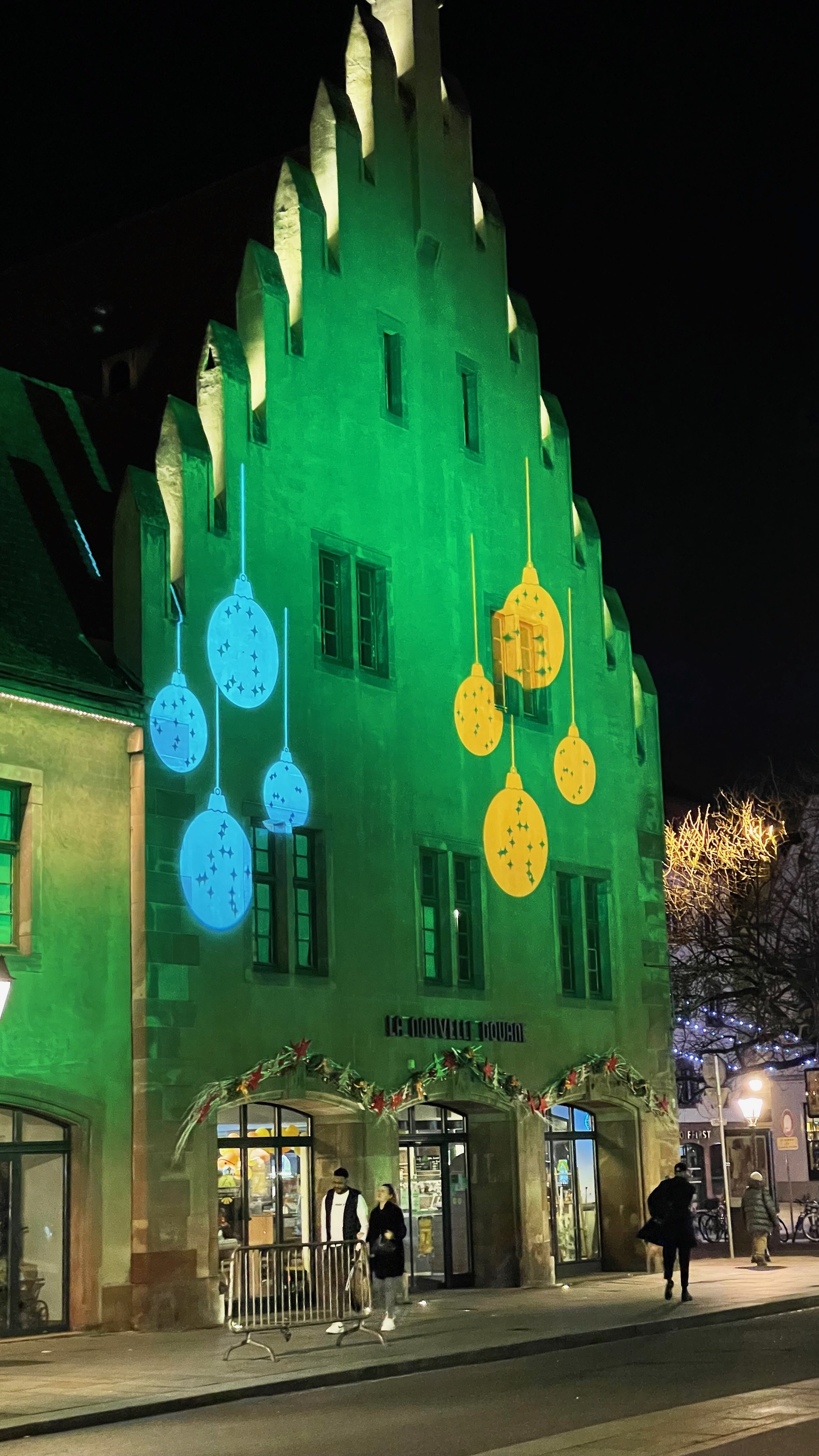 Building facade lit with green light and festive decorations at night, featuring projected images of Christmas ornaments in blue and yellow, with people walking nearby.