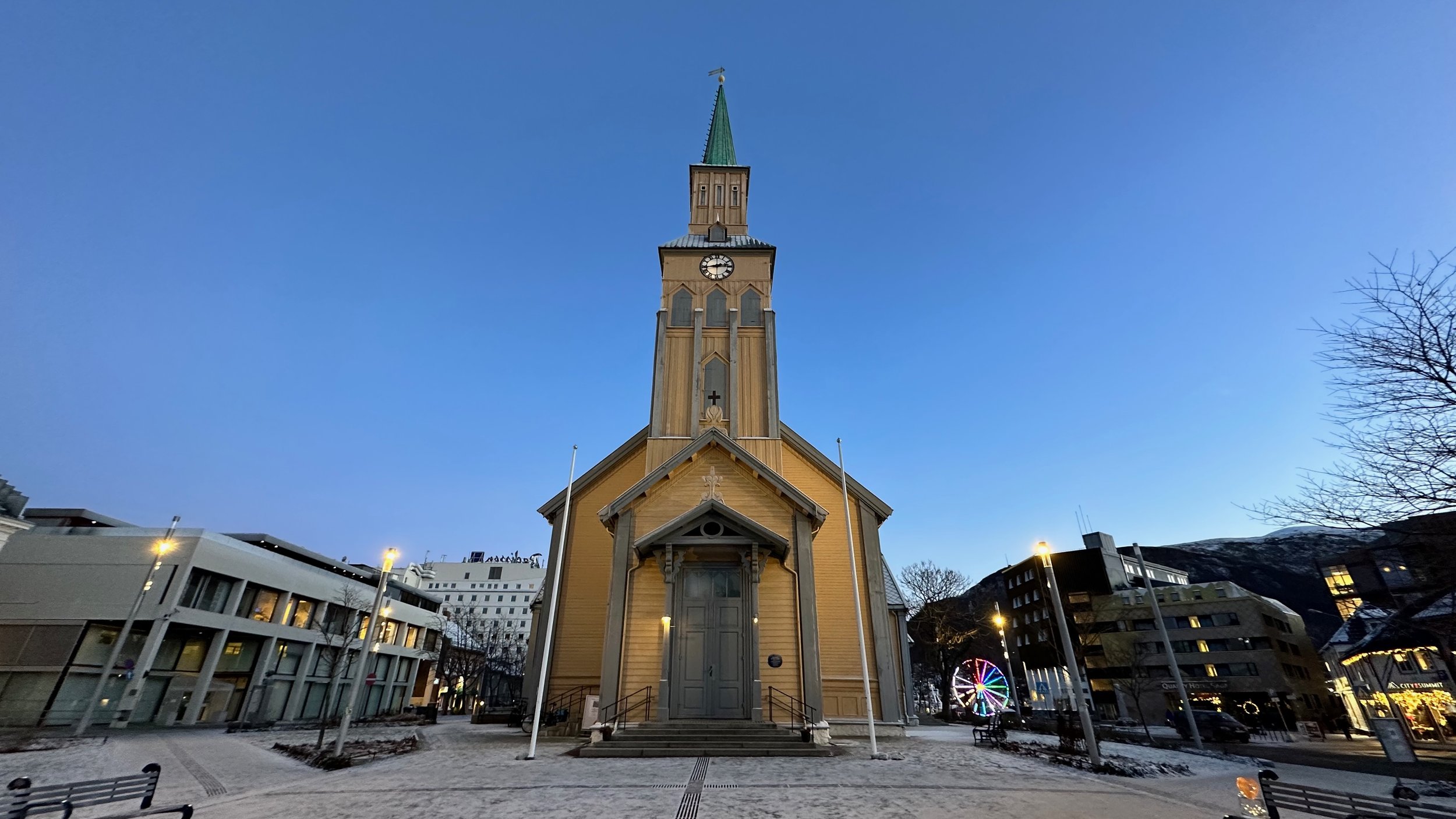 A historic wooden church with a tall spire and clock, surrounded by modern buildings and bare trees on a winter evening. A Ferris wheel is visible in the background.
