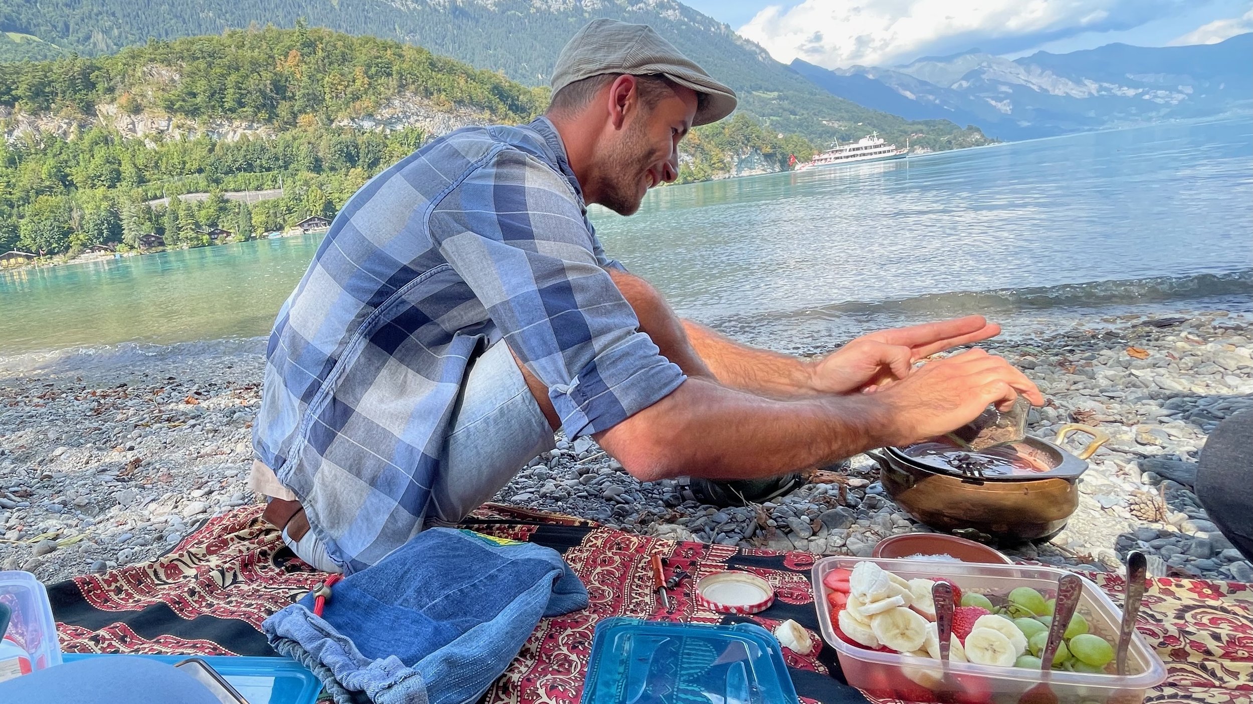 Man sitting by a lake preparing a meal on a picnic blanket with fruit and pot, scenic mountain and water view in the background.