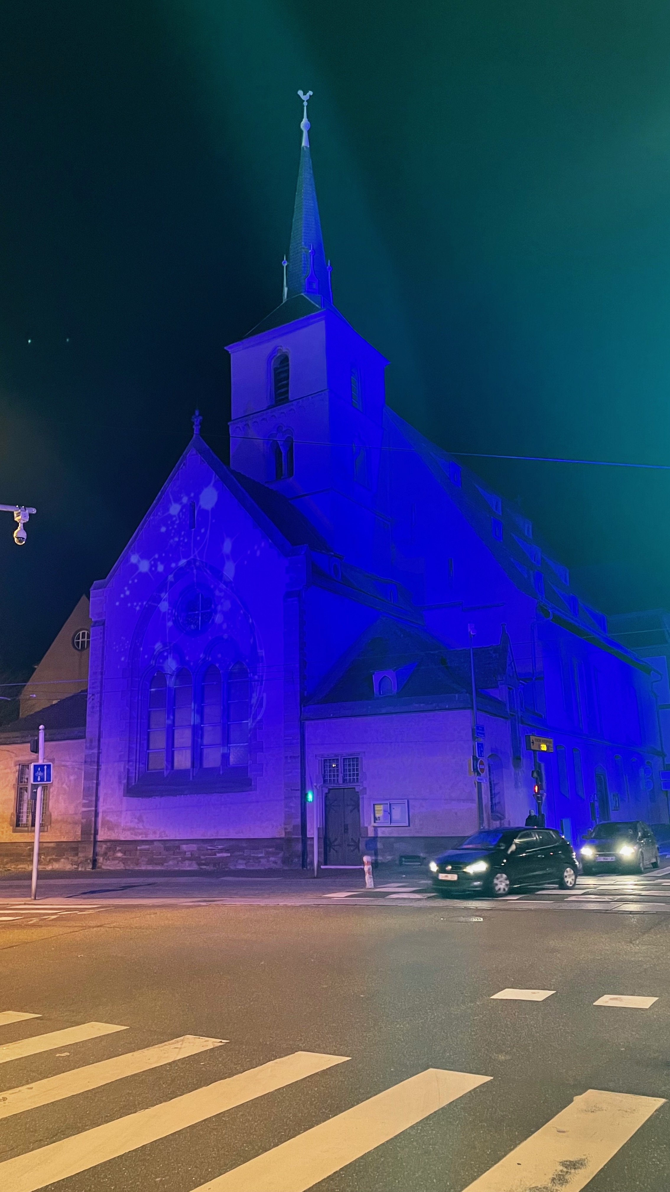 Church building illuminated in blue light at night with cars passing by on the street.