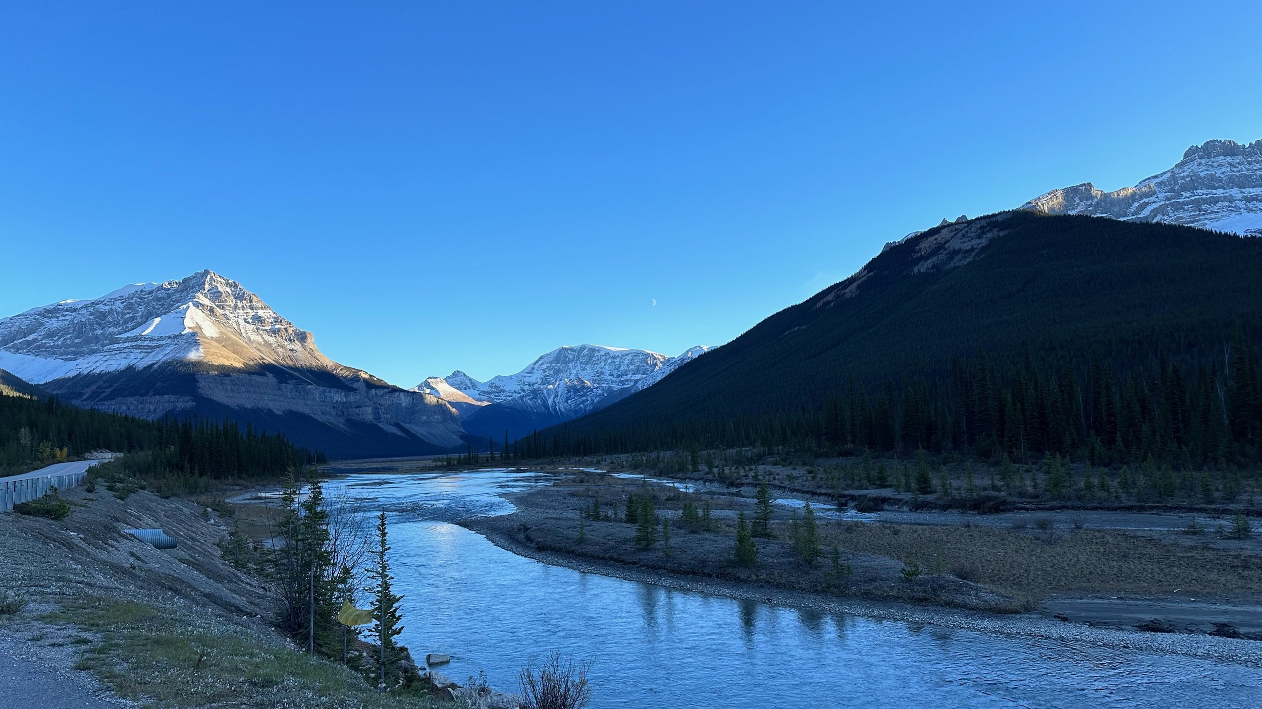 Mountain landscape with a river, snow-capped peaks, pine trees, and a clear blue sky.