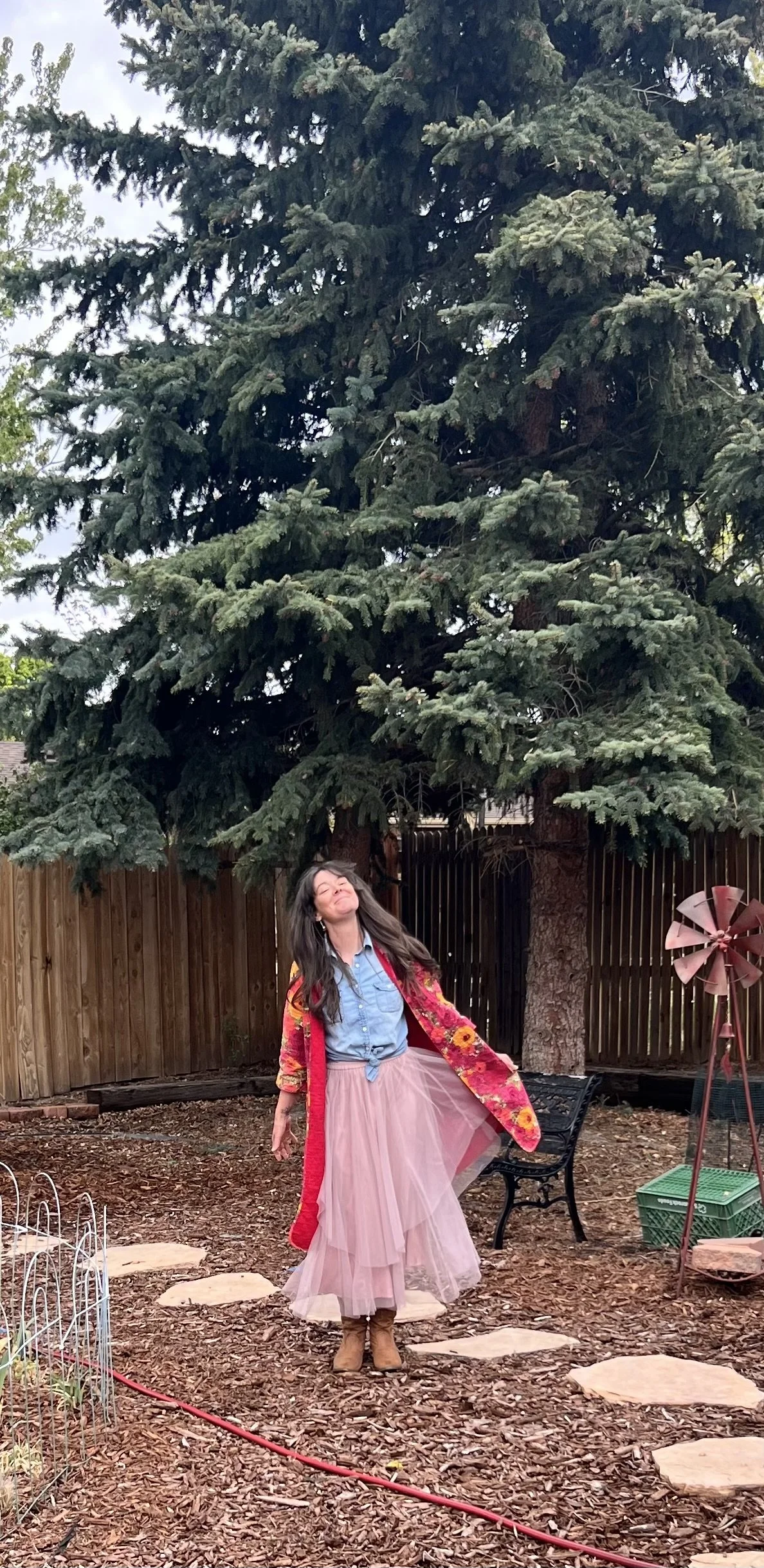 “Woman standing under a large evergreen tree in a backyard, smiling with eyes closed and wearing a colorful floral coat and soft pink skirt”