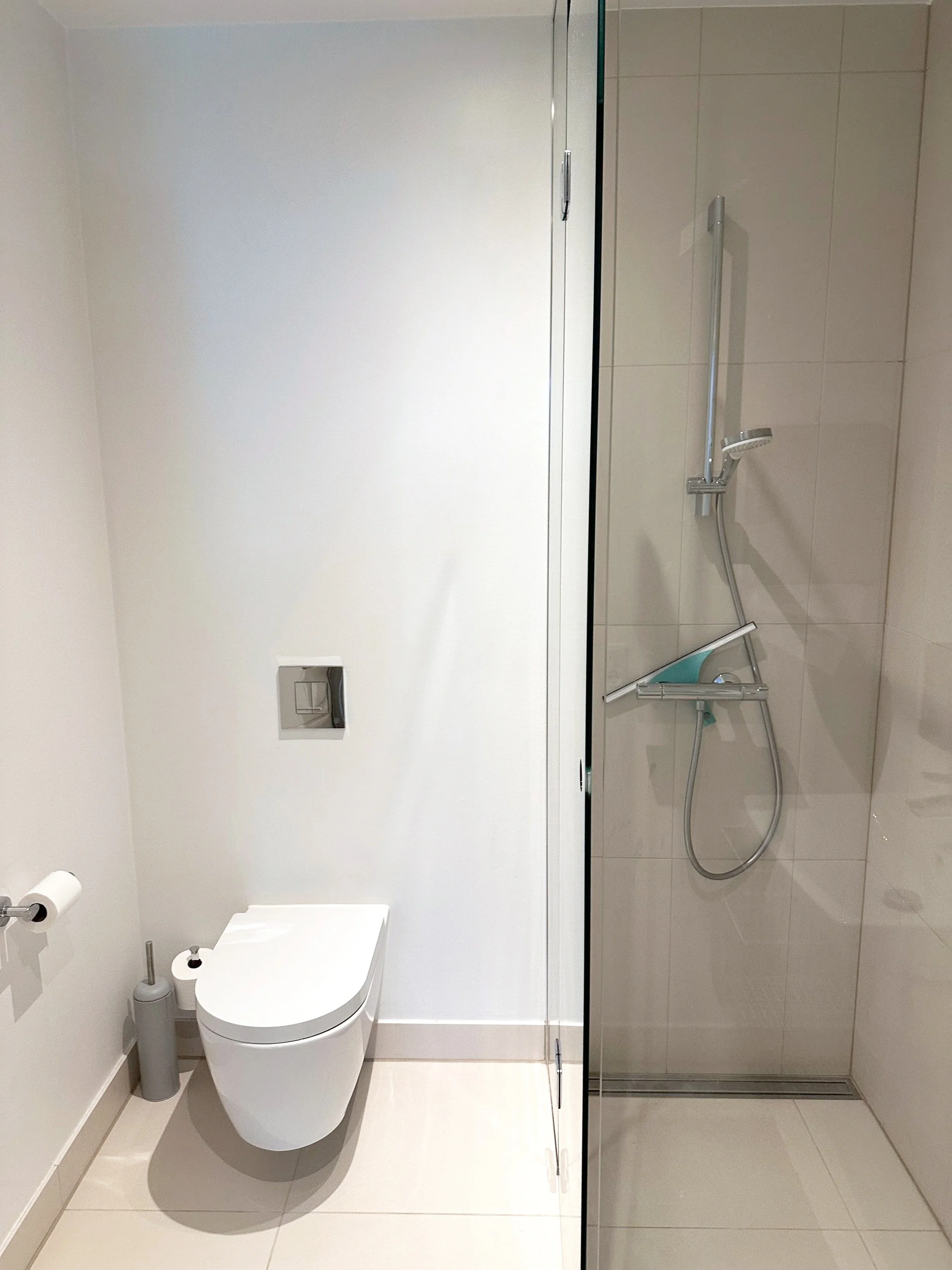 Et moderne badeværelse med toilet og bruser. Toilet er placeret mod venstre side af billedet, med en toiletbørste og toiletpapirholder ved siden af. Bruseren er i højre del af billedet med et vægmonteret bruserhoved og glasvæg.