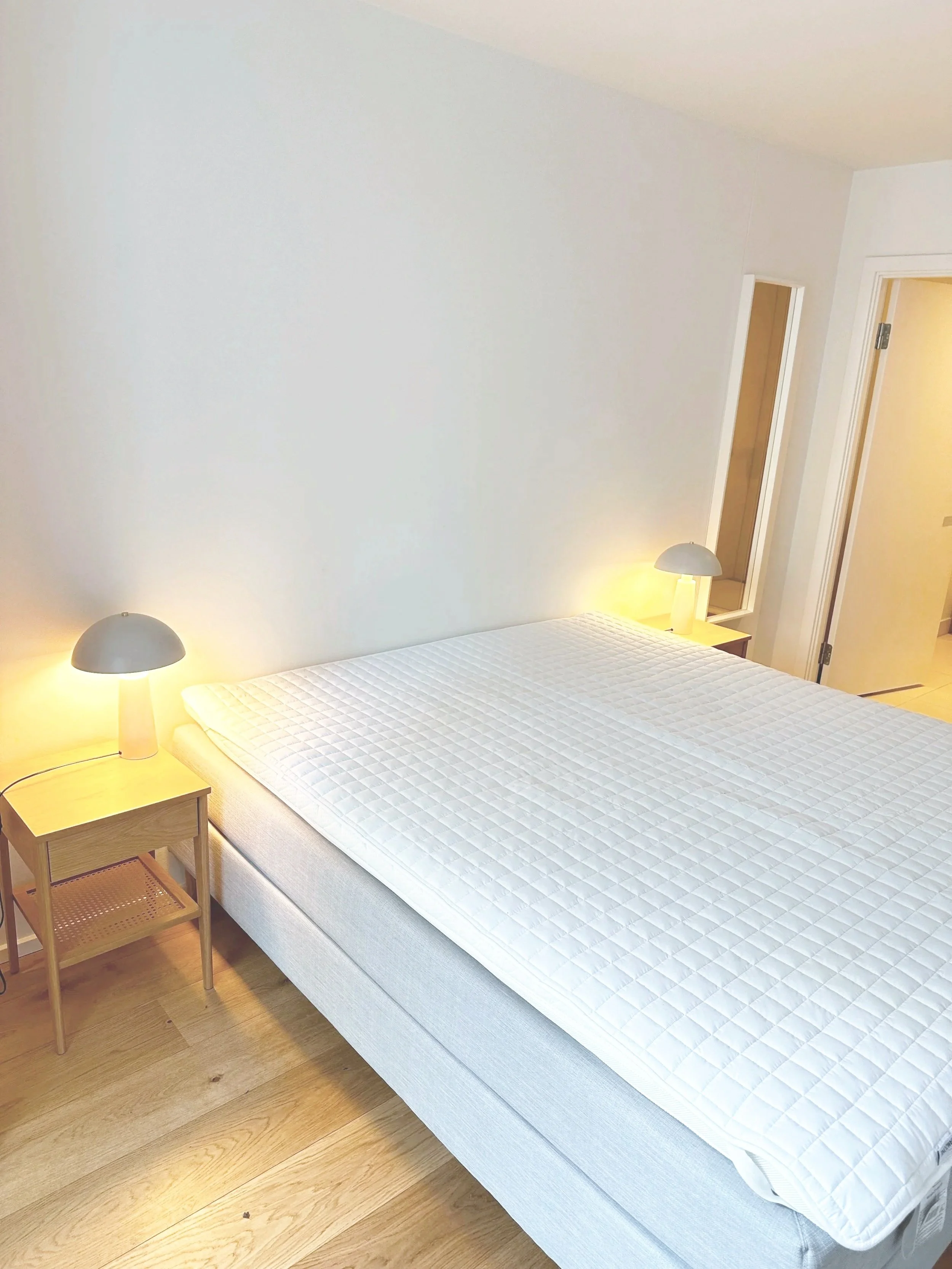 Et soveværelse med en stor seng, der har en hvid mattress og to små sideborde med lamper, lyse trægulve, hvid væg og en dør med spejl i rammen.