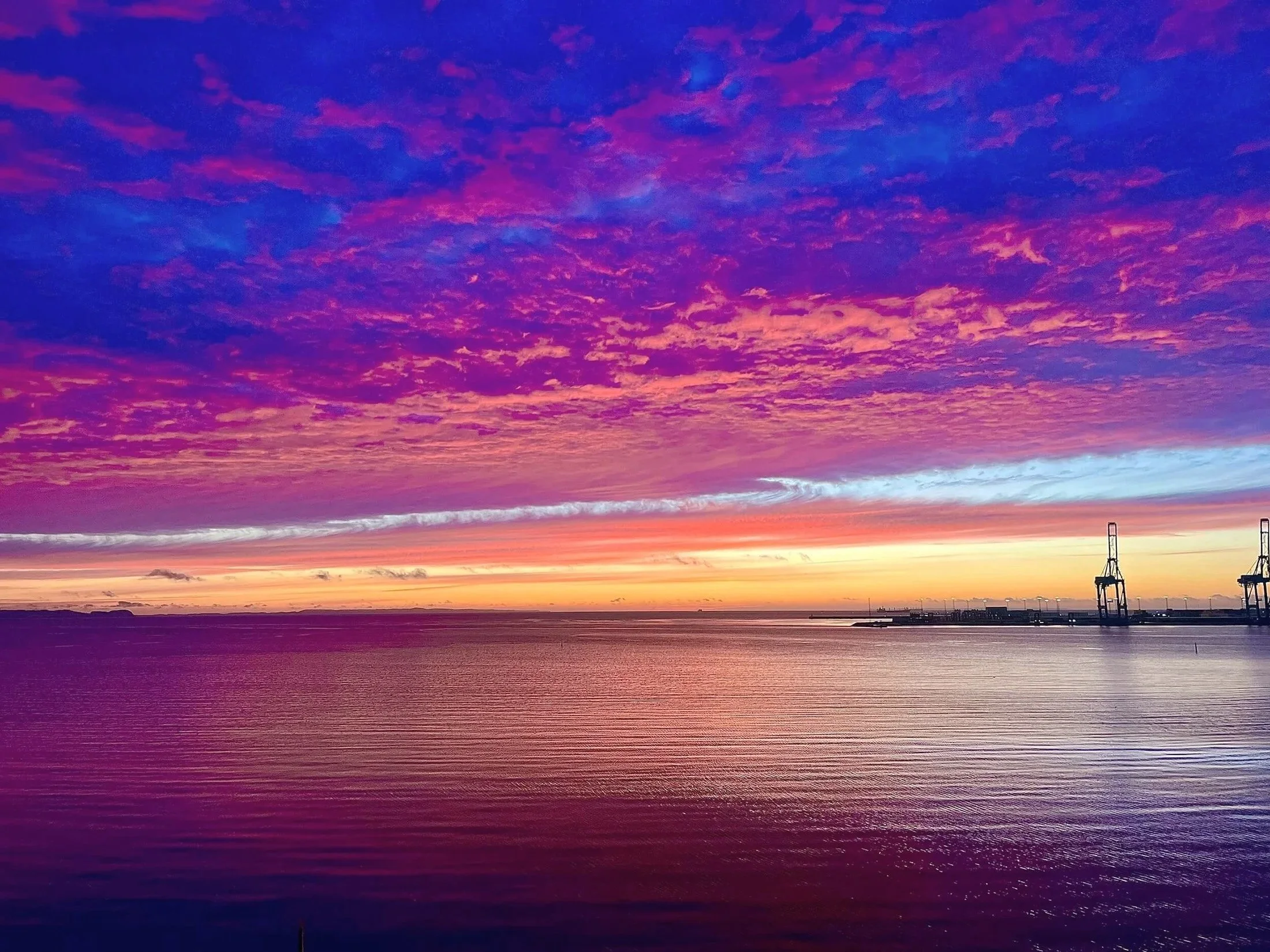 Farvet himmel ved solnedgang med rosa, lilla og orange nuancer over havet, med silhuetter af broer i horisonten.