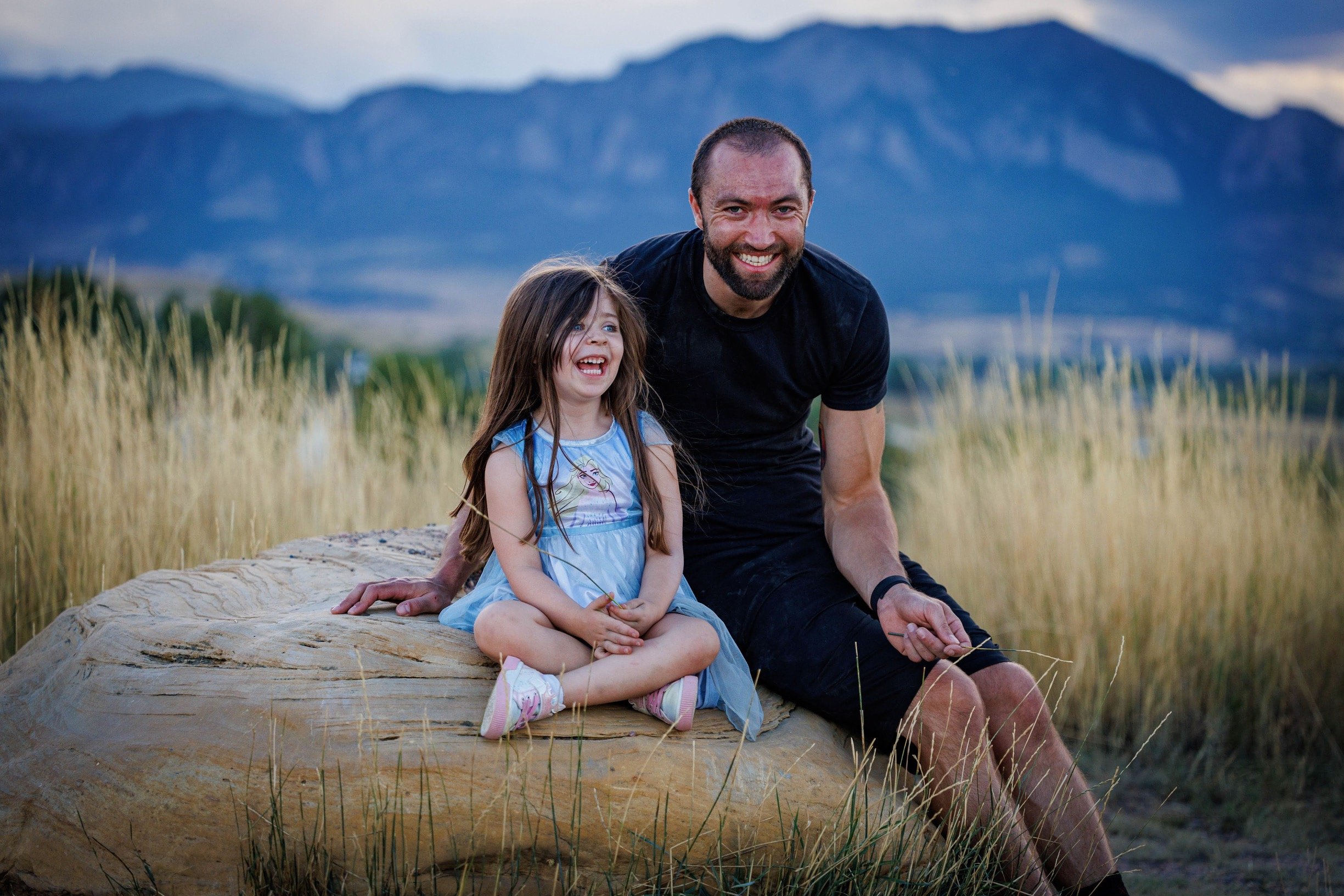 better everyday adaptive fitness coach james anderson wearing black shirt sitting on a rock with daughter wearing elsa dress smiling and laughing together in a mountain area