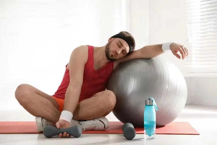 Man showing signs of exhaustion from a workout with free weights, water, and workout ball