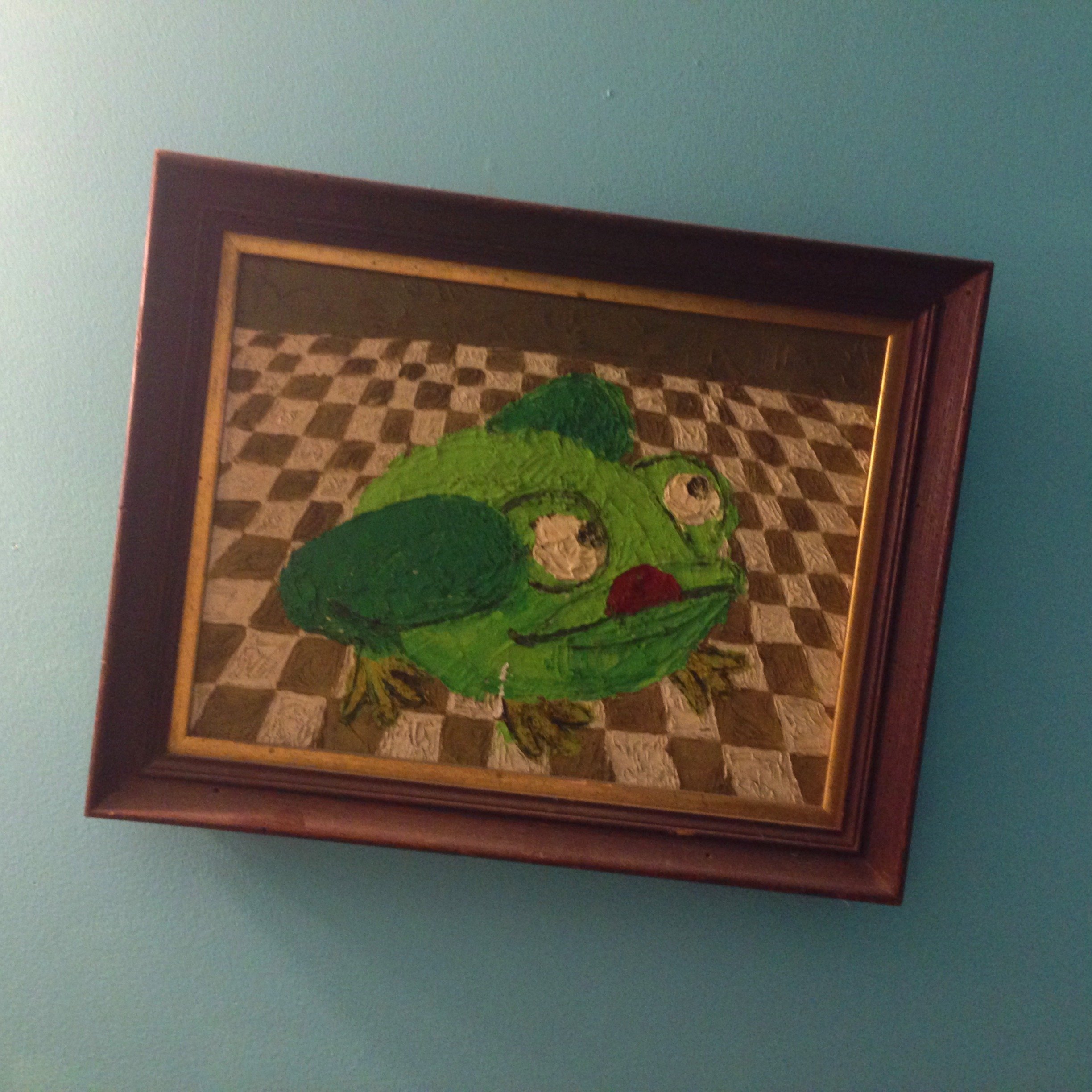 Whimsical painting of a green frog with large eyes on a checkered floor, in a wooden frame, against a blue wall.