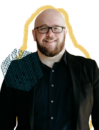 Portrait of a smiling man with glasses, a beard, wearing a dark shirt and jacket, standing against a colorful abstract background.