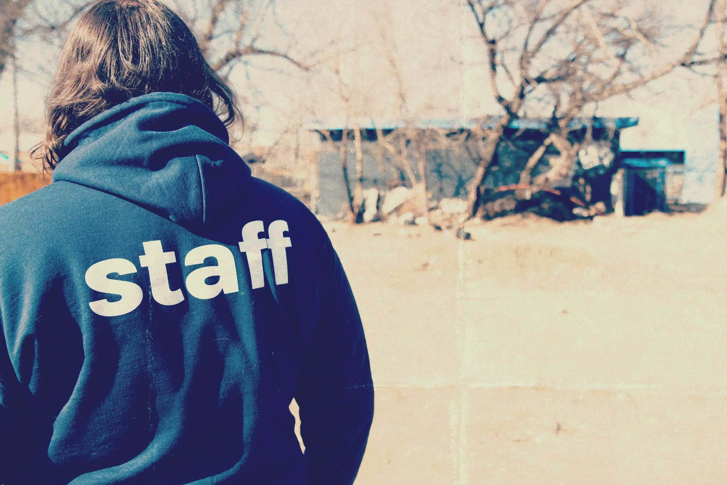 Person wearing a navy hoodie with the word 'staff' printed on the back, standing outdoors near a chain-link fence and leafless trees in the background.