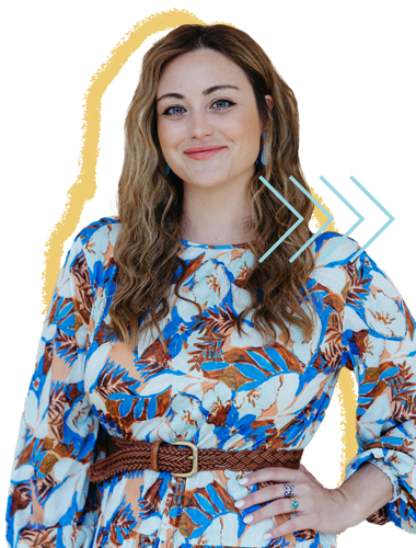 A woman with wavy brown hair and blue eyes, smiling and wearing a floral dress with shades of blue, brown, and cream, standing with one hand on her hip.