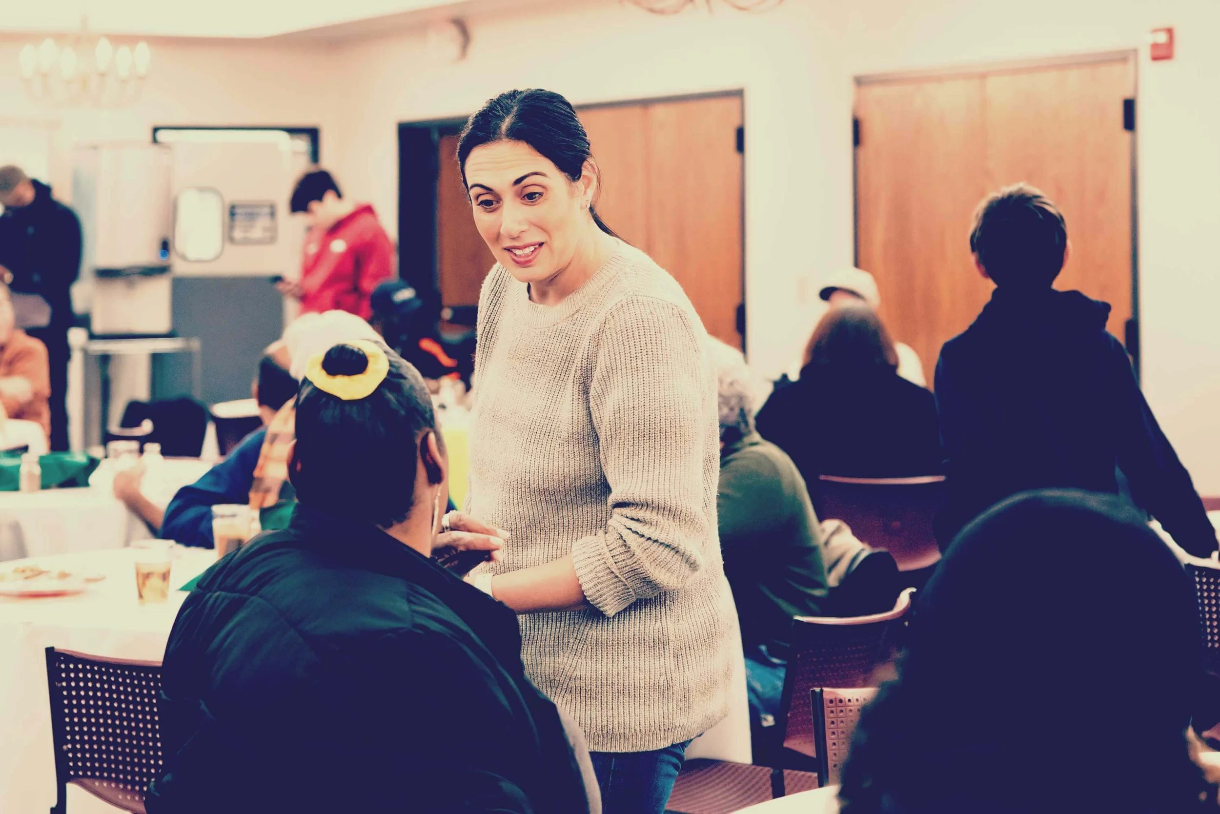 A woman in a beige sweater talking to a woman with a yellow hair accessory in a busy indoor setting with people seated at tables.