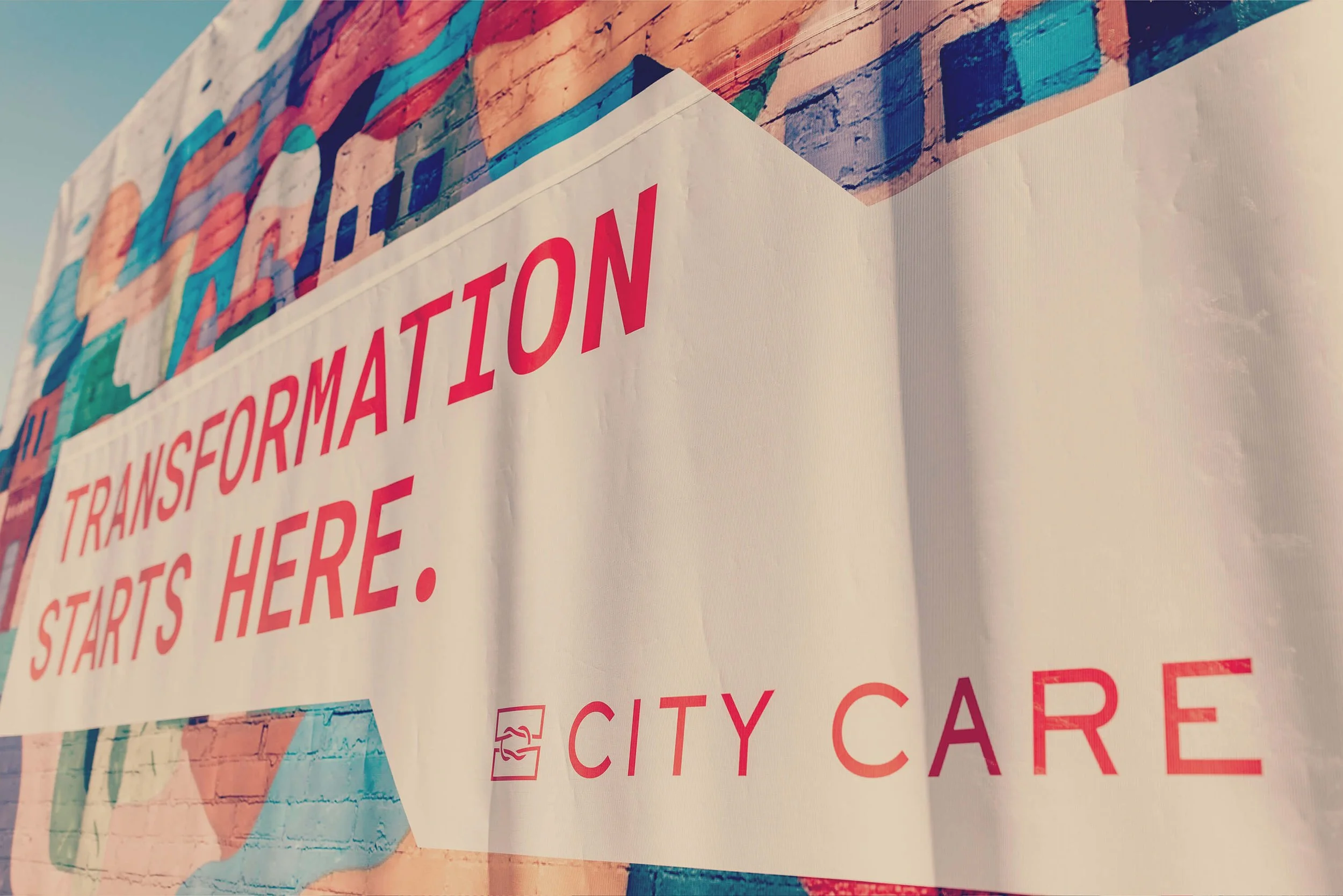 A banner that reads "Transformation Starts Here" with the logo and name "City Care" at the bottom, displayed outdoors against a brick wall.