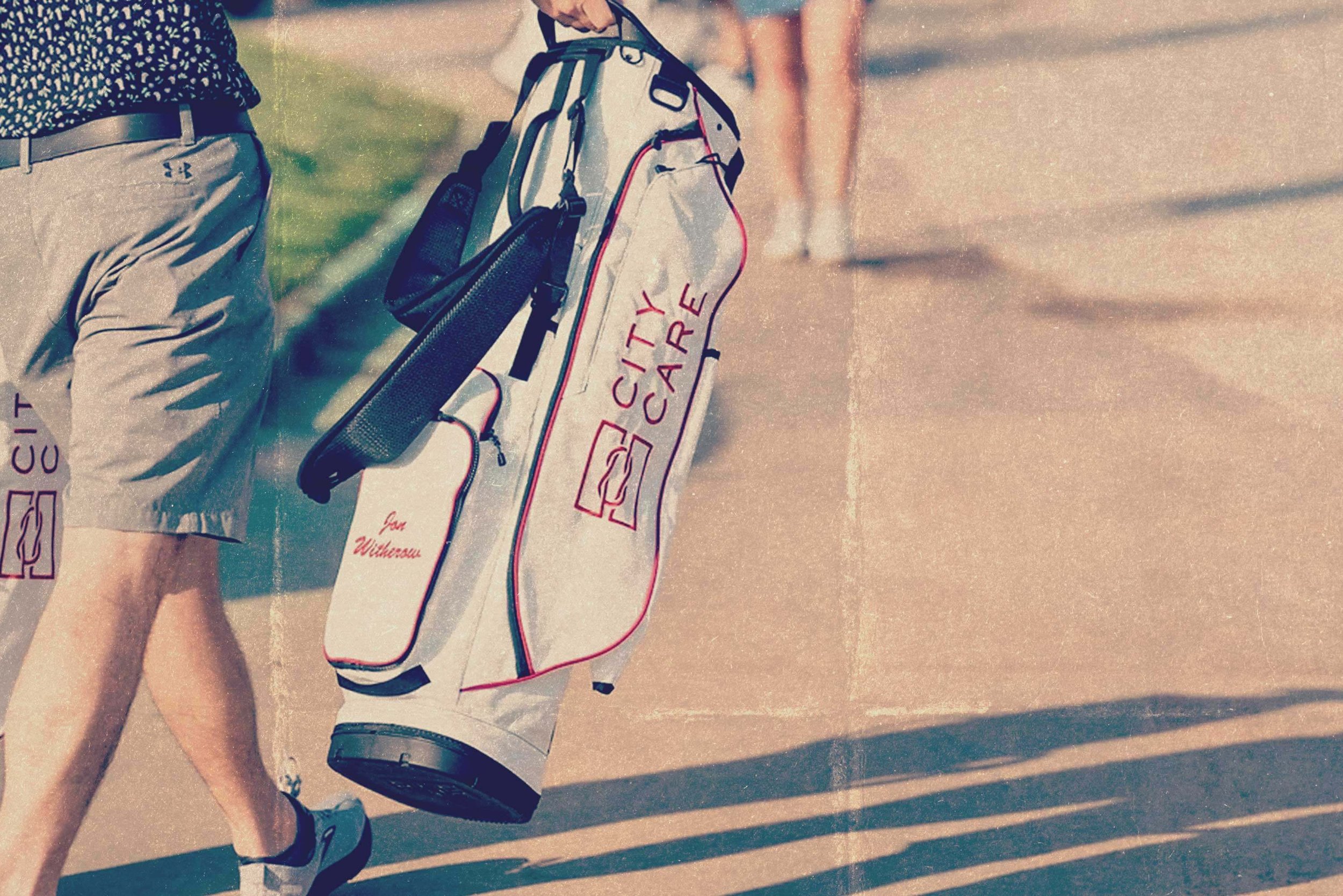 A person walking on a sidewalk, carrying a golf bag with clubs, with their left arm and part of their legs visible. The golf bag is labeled "City Care" and has a smaller bag attached to it. The person is wearing shorts and sneakers.