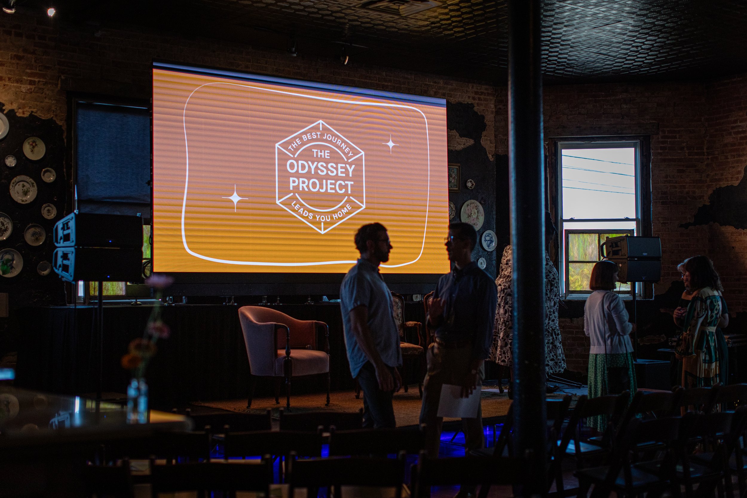 People silhouetted against a large screen displaying 'The Odyssey Project' logo at an indoor event.