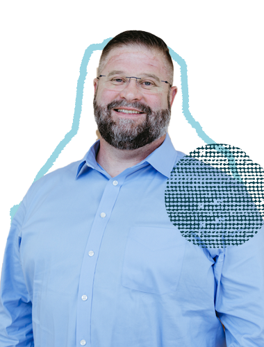 A smiling man with glasses and a beard wearing a light blue dress shirt, standing against a light background.